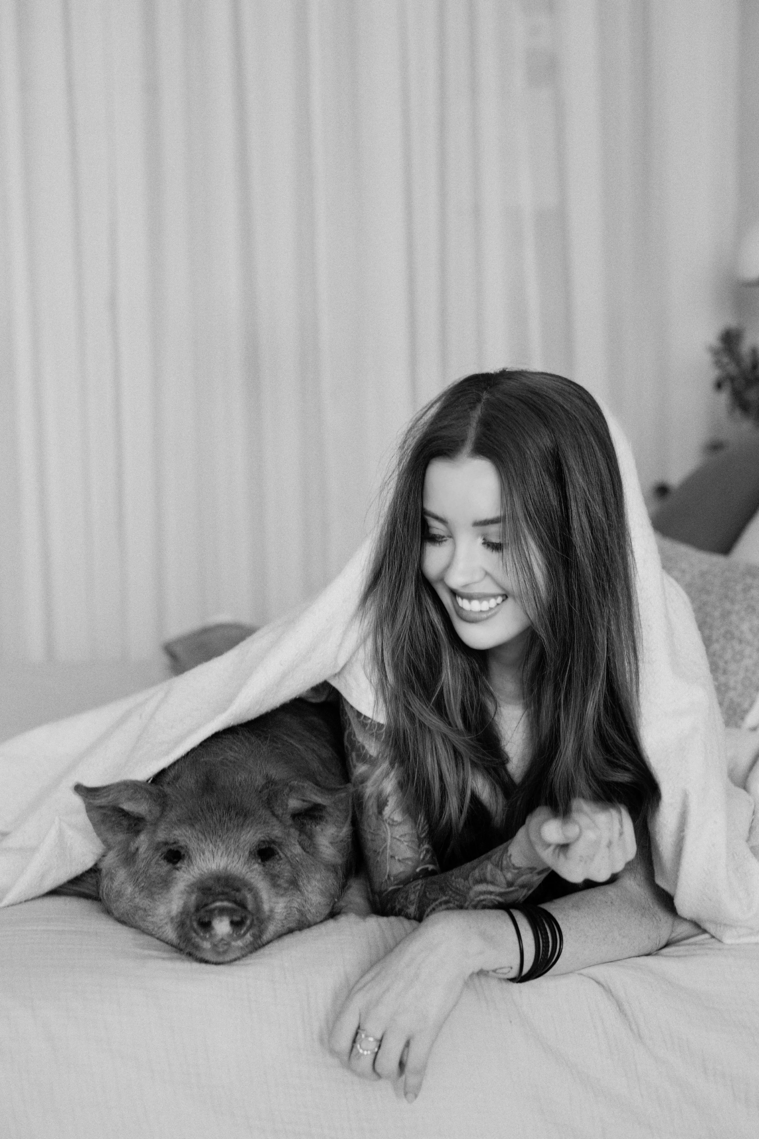 A woman and a pig lying on a bed, smiling, with a blanket draped over them.