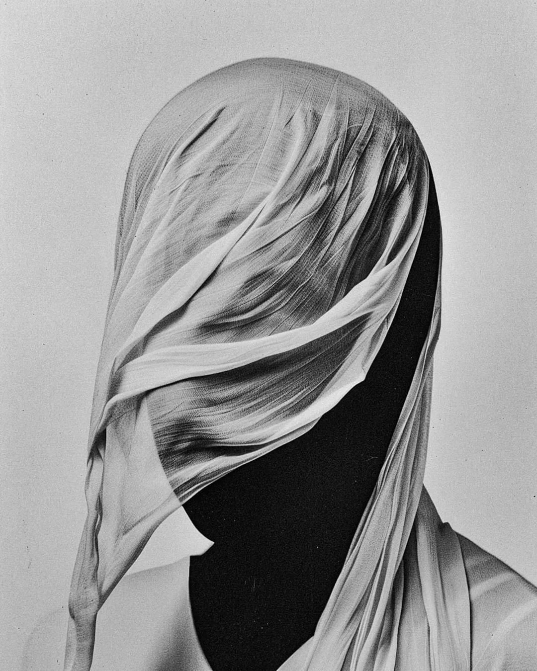 Black and white photograph of a human figure with a head wrapped in layered, flowing fabric that obscures the face, against a plain background.