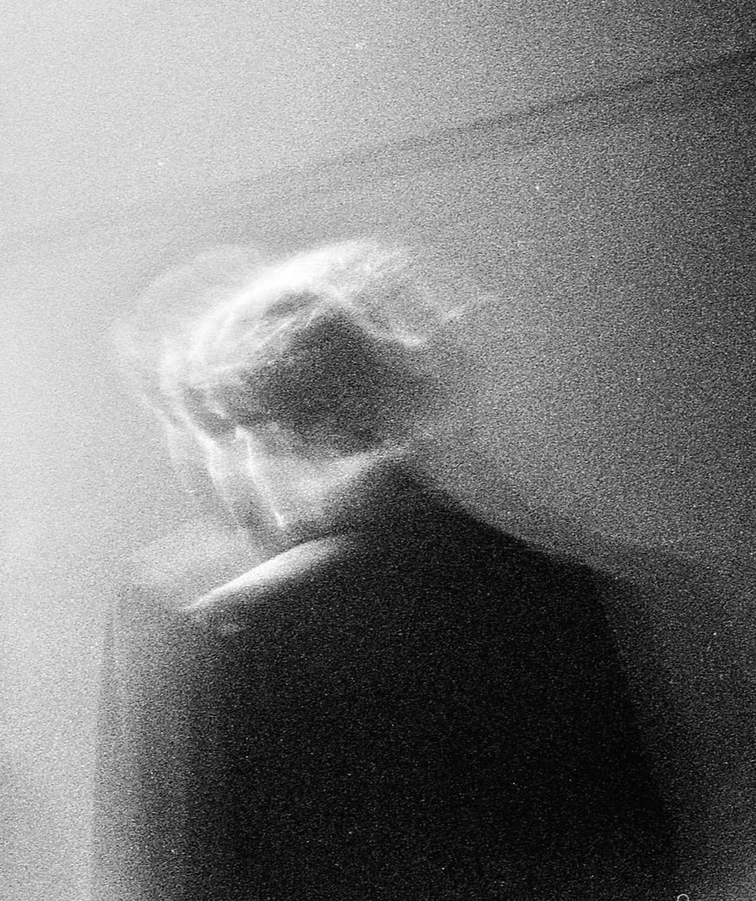 Black and white photo of a person looking through a textured glass or frosted window, with only their profile visible and their eye partially obscured. They are resting their chin on their hand.