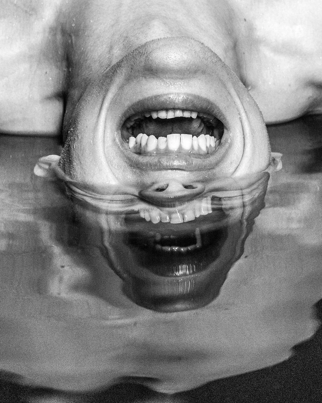 A person with a big smile, head upside down, floating in water, showing their teeth, with their reflection visible in the water.