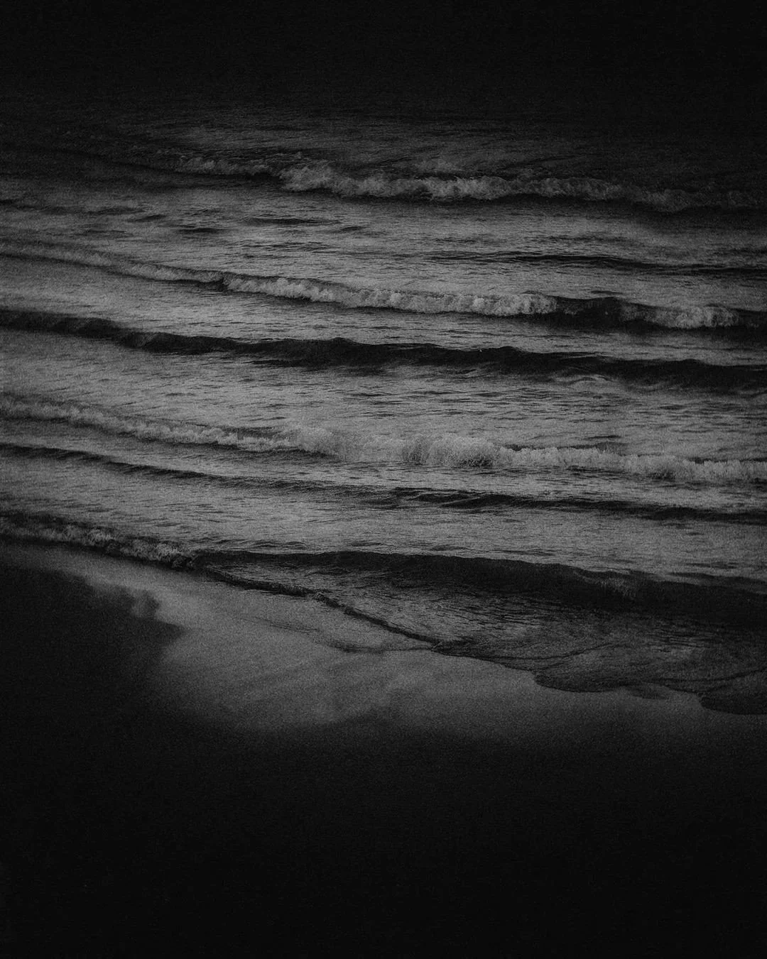 Black and white photo of ocean waves gently hitting the shore at night.