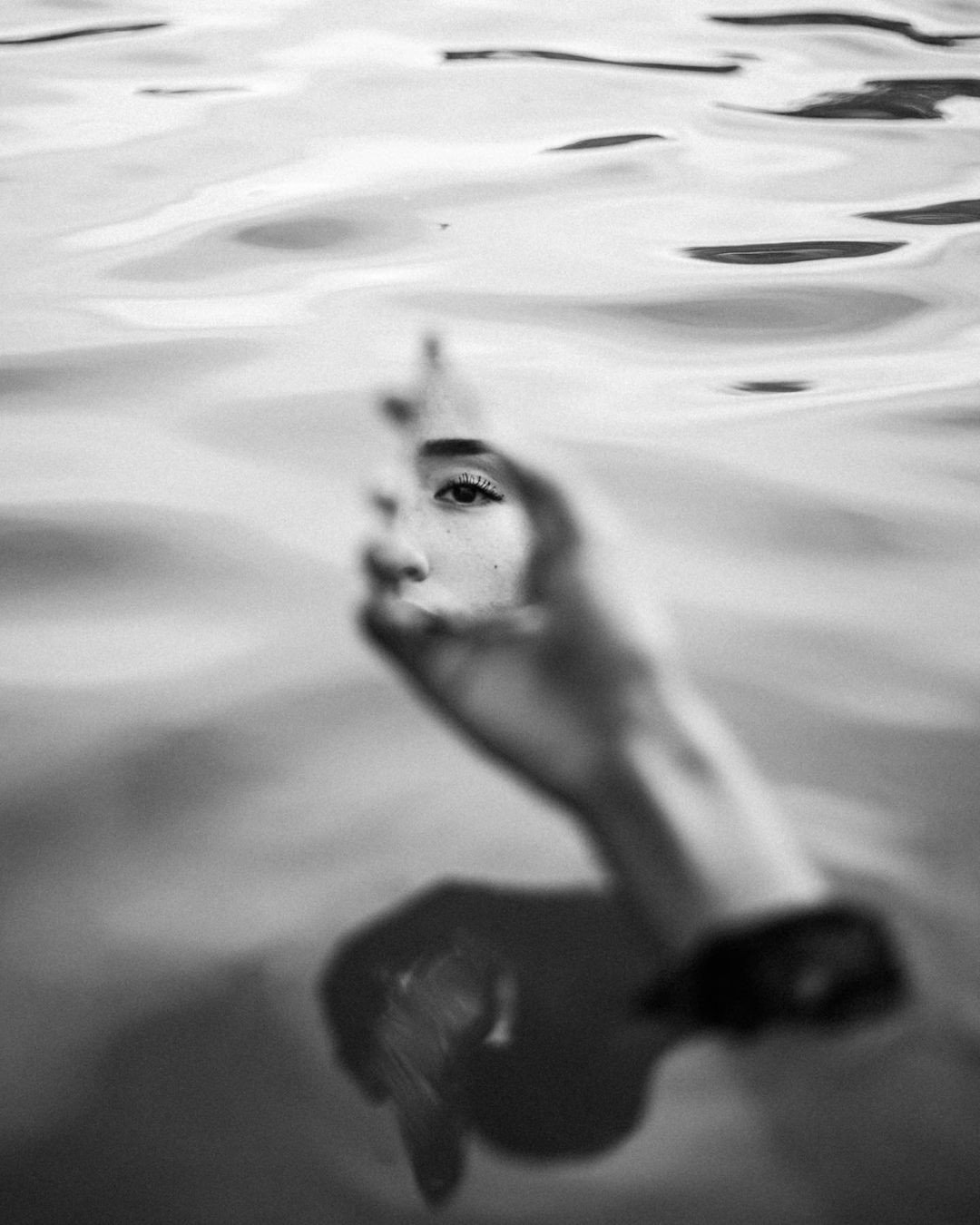 A black and white photograph of a person's face and hand visible through water, with the person's eye staring directly at the camera as they hold up their hand with fingers curved, swimming in water with visible ripples.