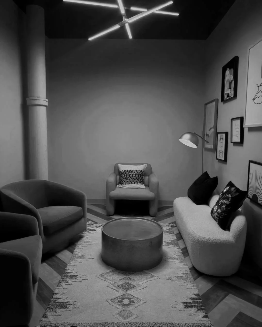 A cozy living room with modern furniture, including a white sofa with black pillows, a plush armchair with a cushion, a circular coffee table, and a gallery wall with framed art. A floor lamp and a geometric ceiling light fixture add to the ambiance, with a patterned rug on wooden floors.