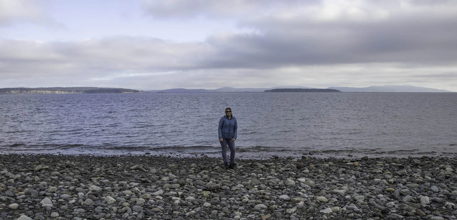 A woman in a blue jacket and sunglasses standing on a rocky beach near a lake with distant islands and a cloudy sky.
