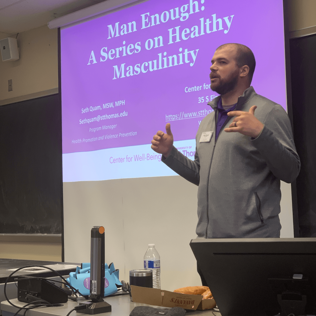 A man in a gray jacket giving a presentation in a classroom, with a slide titled 'Man Enough: A Series on Healthy Masculinity' projected behind him.