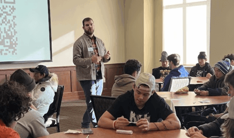 A man standing at the front of a room filled with students, giving a presentation.