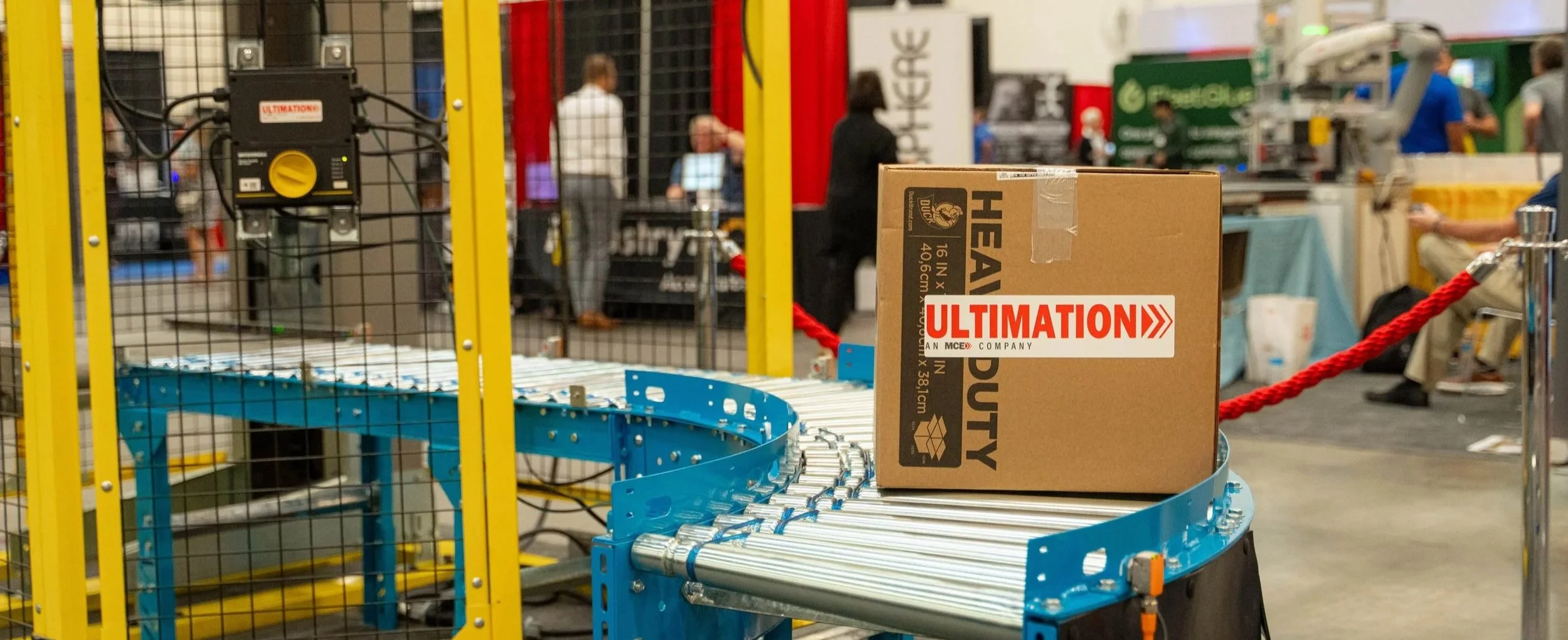 A cardboard box labeled 'Ultimate' on a blue conveyor belt at a trade show or exhibition. In the background, there are people walking and standing around booths with displays, including one for 'Hef'. The conveyor belt is part of a machinery setup enclosed in a yellow frame with black wire mesh.