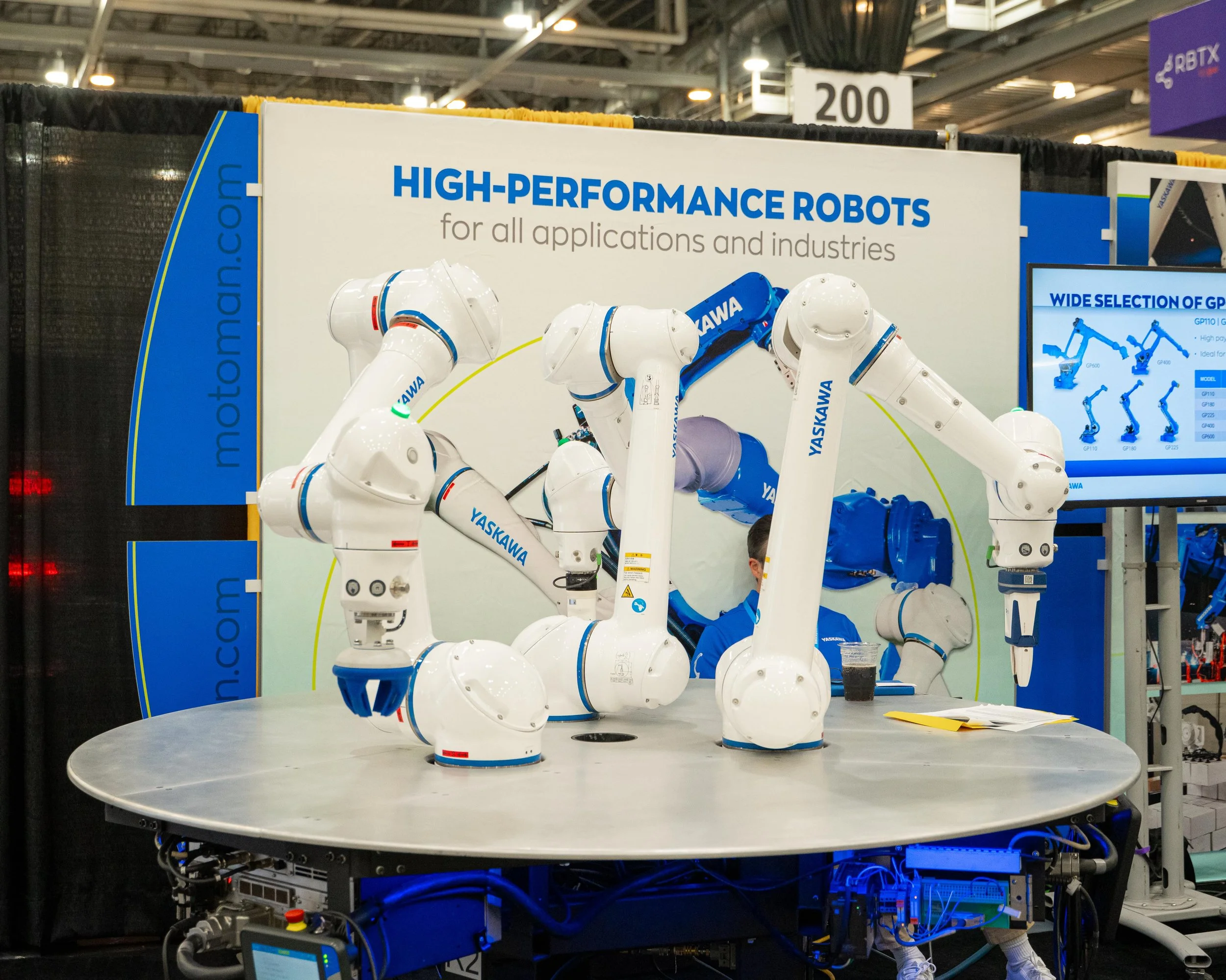 A demonstration of high-performance industrial robots is shown with multiple robotic arms arranged around a circular table. The robots are branded 'YASKAWA' and are displayed at an exhibition or trade show, with a large sign in the background advertising their applications in various industries.