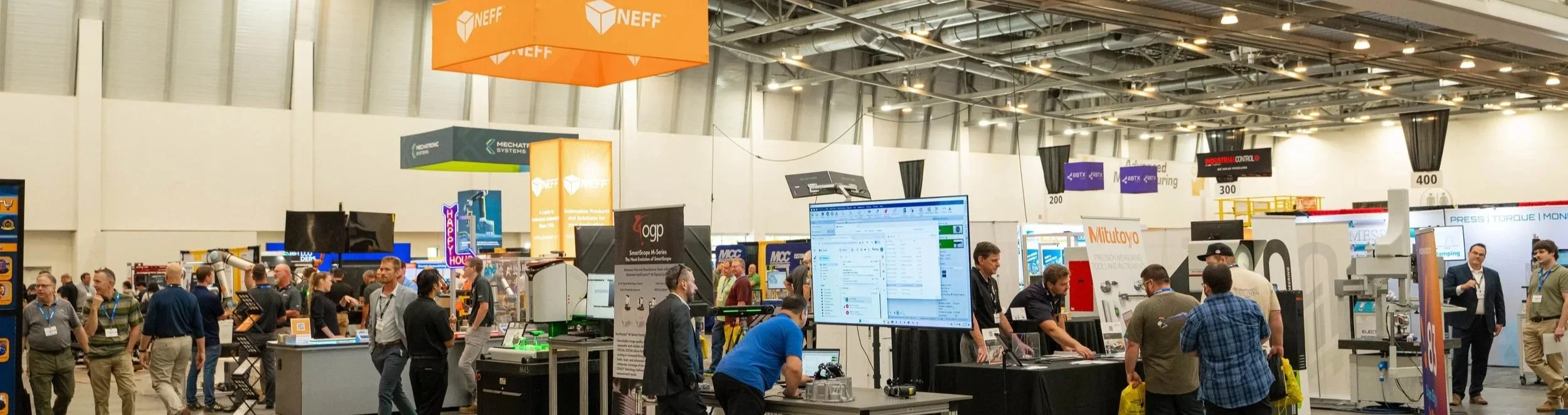 A busy trade show or conference with various booths and people networking. Large screens display data and presentations, and there are banners with company logos such as NEFF and Mitutoyo.
