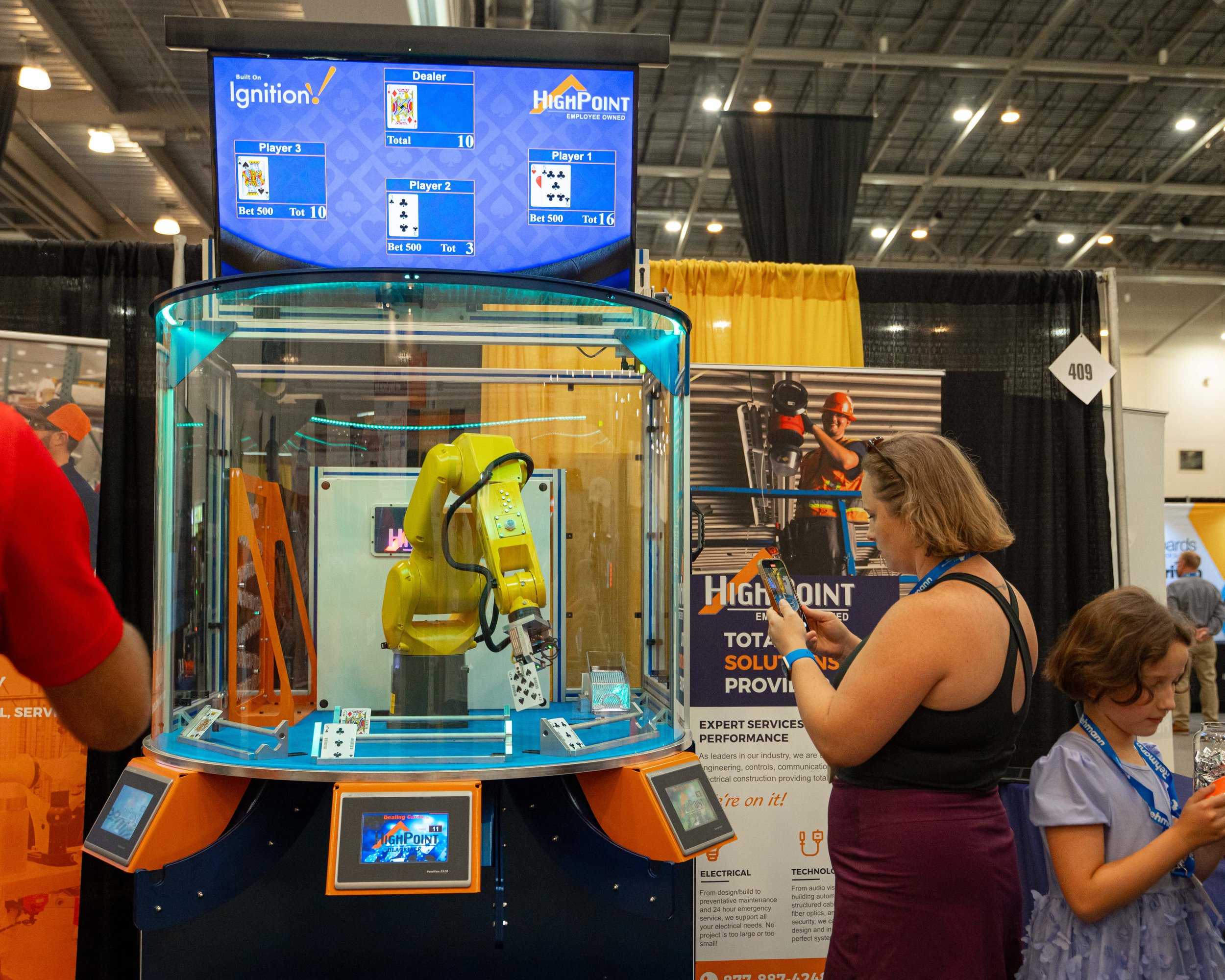 A robotic arm deals playing cards inside a glass enclosure at a trade show, with a woman and a girl observing and using a smartphone nearby. A large monitor displays digital cards and game results.