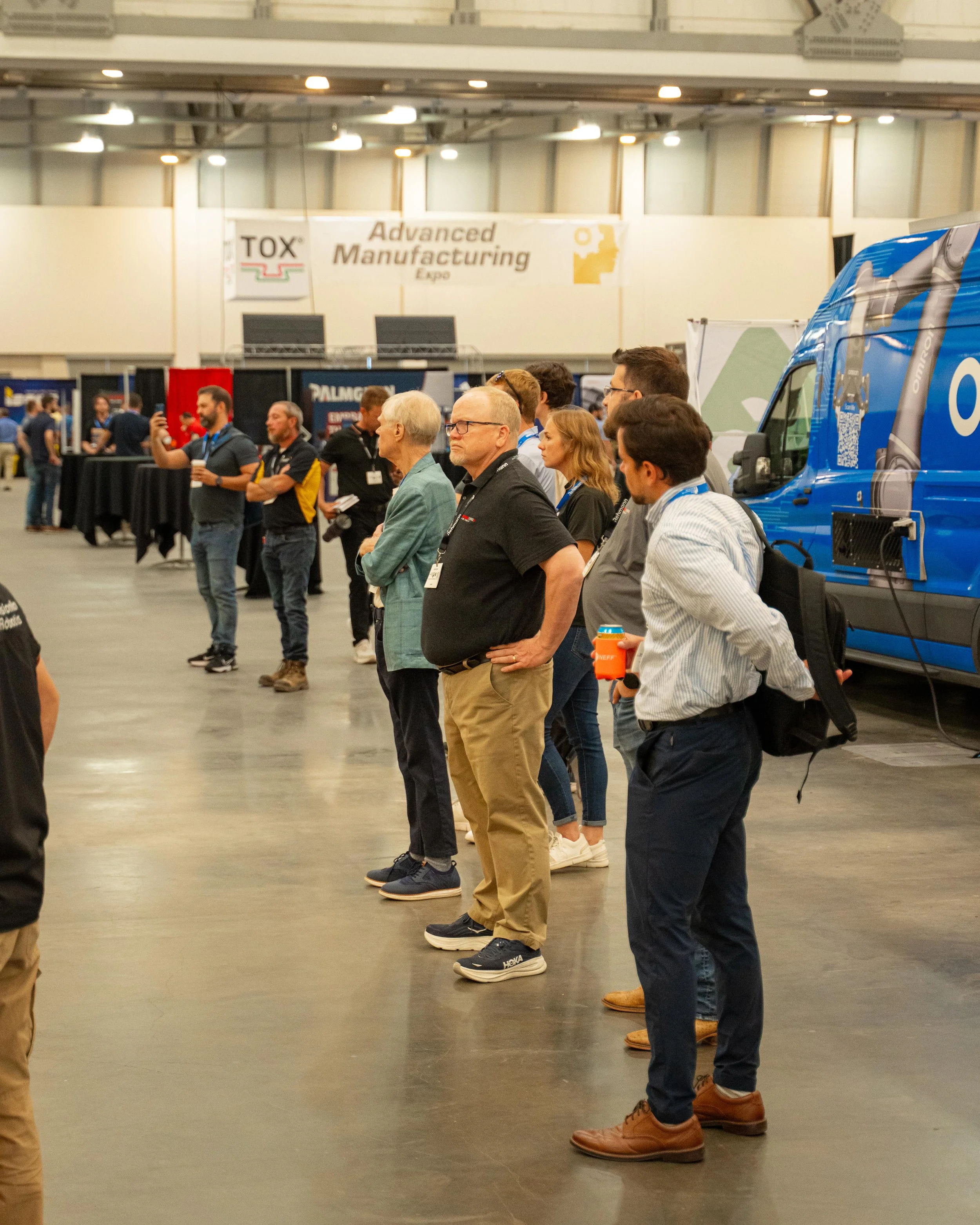 People attending an advanced manufacturing expo, standing in a line inside a large exhibition hall with booths and a blue vehicle on the side.