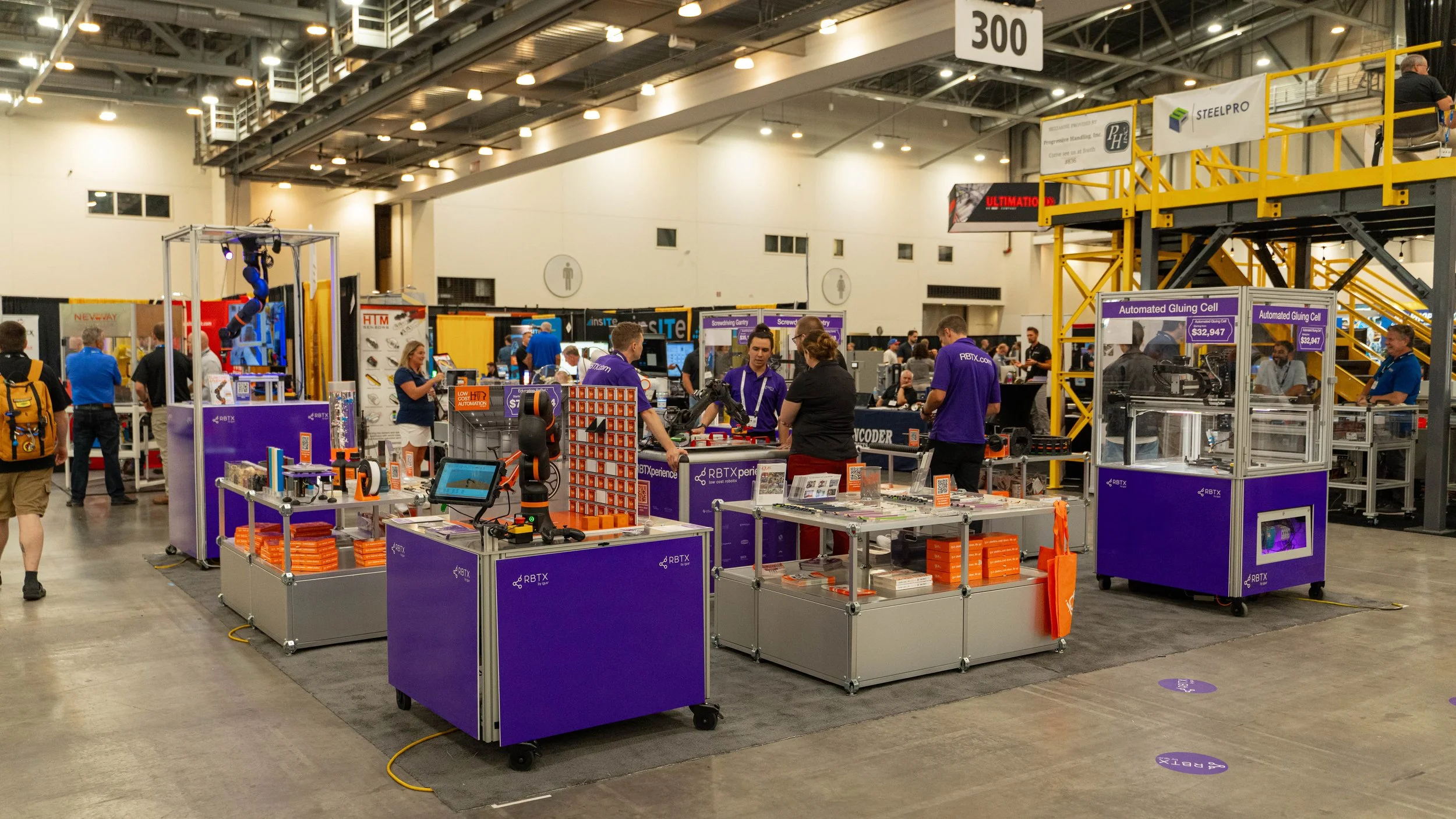 Trade show booth with robotic and automation equipment, people interacting, purple display counters, and yellow staircase in a large exhibition hall.