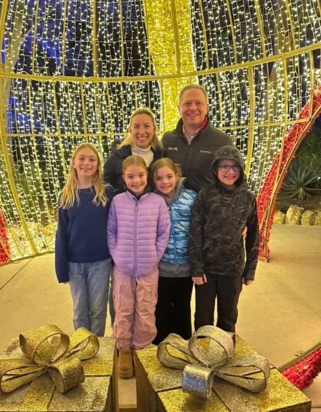 Family of six smiling in front of a festive holiday light display with large gold gift boxes in the foreground.