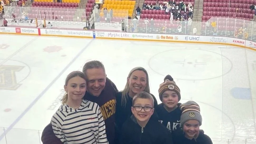 A family of five posing in front of a hockey rink, with the ice and spectators in the background.