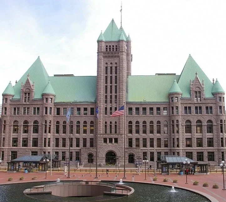 Historic courthouse with a central tower, green roof, and flags in front, surrounded by a brick plaza and water fountain.