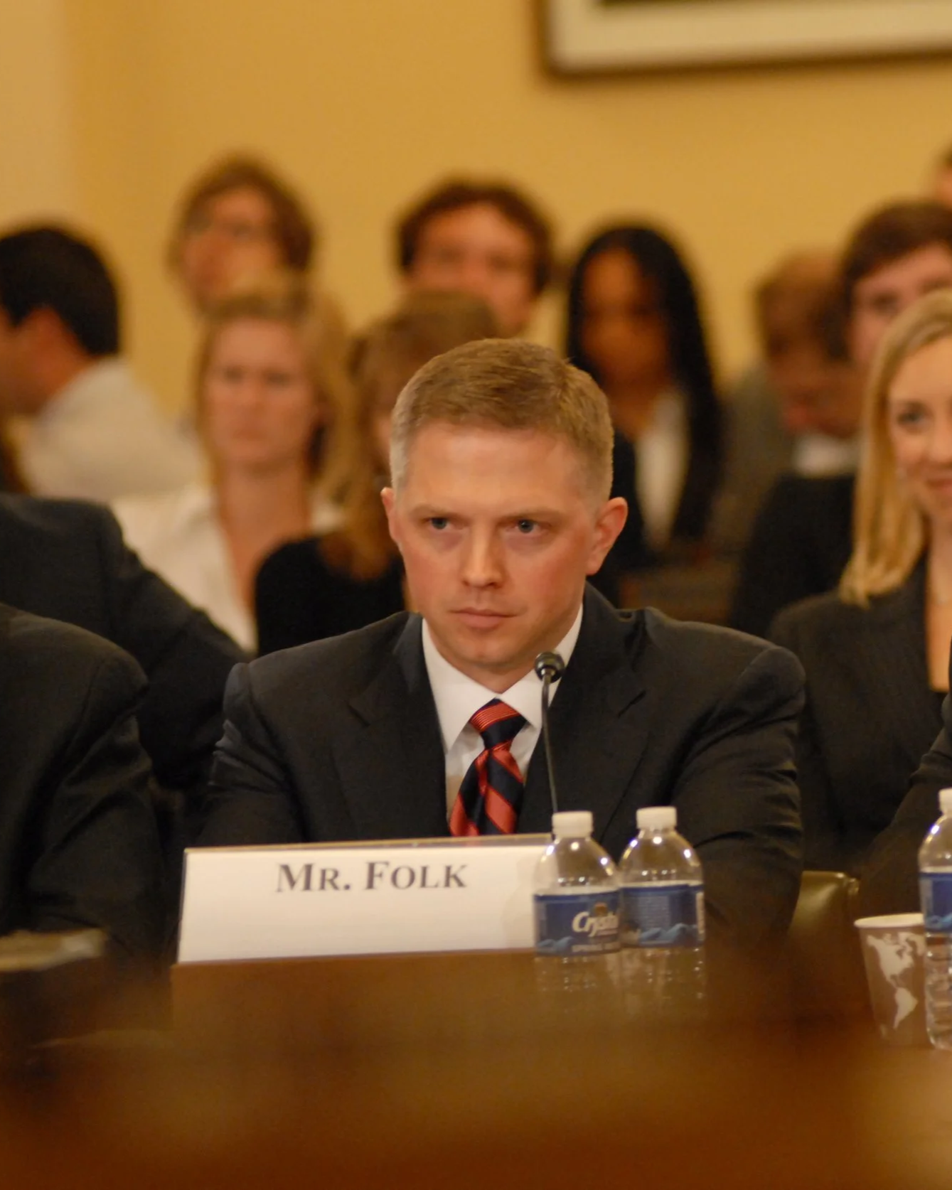 A man in a suit and tie sitting at a table with a nameplate that reads 'Mr. Folk', surrounded by other people in a conference or meeting room.