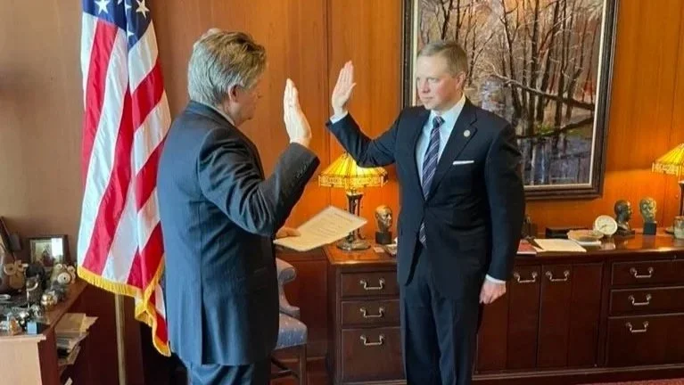 Two men in suits giving each other a high five in an office with an American flag, wooden wall, and framed picture.