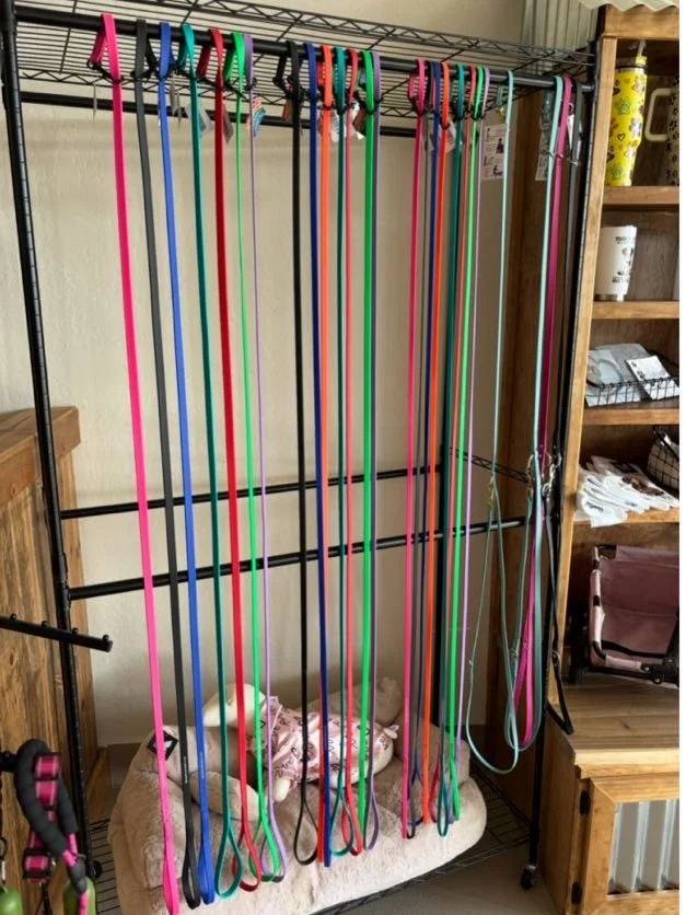 Colorful resistance bands hanging on a black metal rack in a home gym or living space, with a small plush dog toy on a beige cushion at the bottom of the rack and wooden shelving on the side.