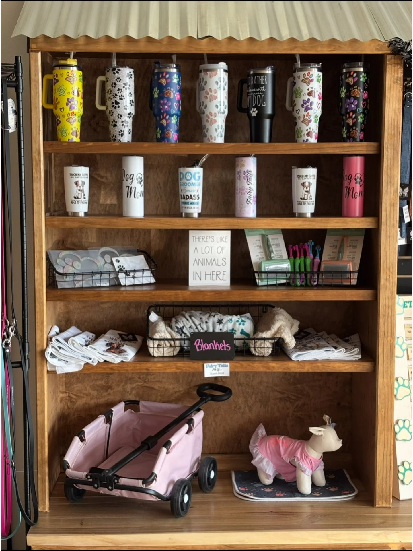 Wooden shelf containing pet and dog-related items, including decorated tumblers on the top shelf, cups with dog-themed sayings on the second shelf, a box of various items, a sign about animals, baskets of blankets and dog accessories, and a pink wagon and plush dog dressed in pink on the bottom shelf.