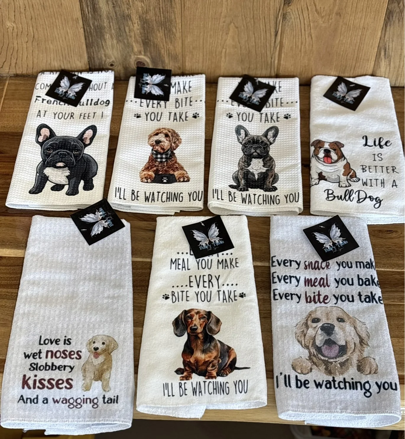 Seven dish towels with dog-themed designs and quotes displayed on a wooden surface.