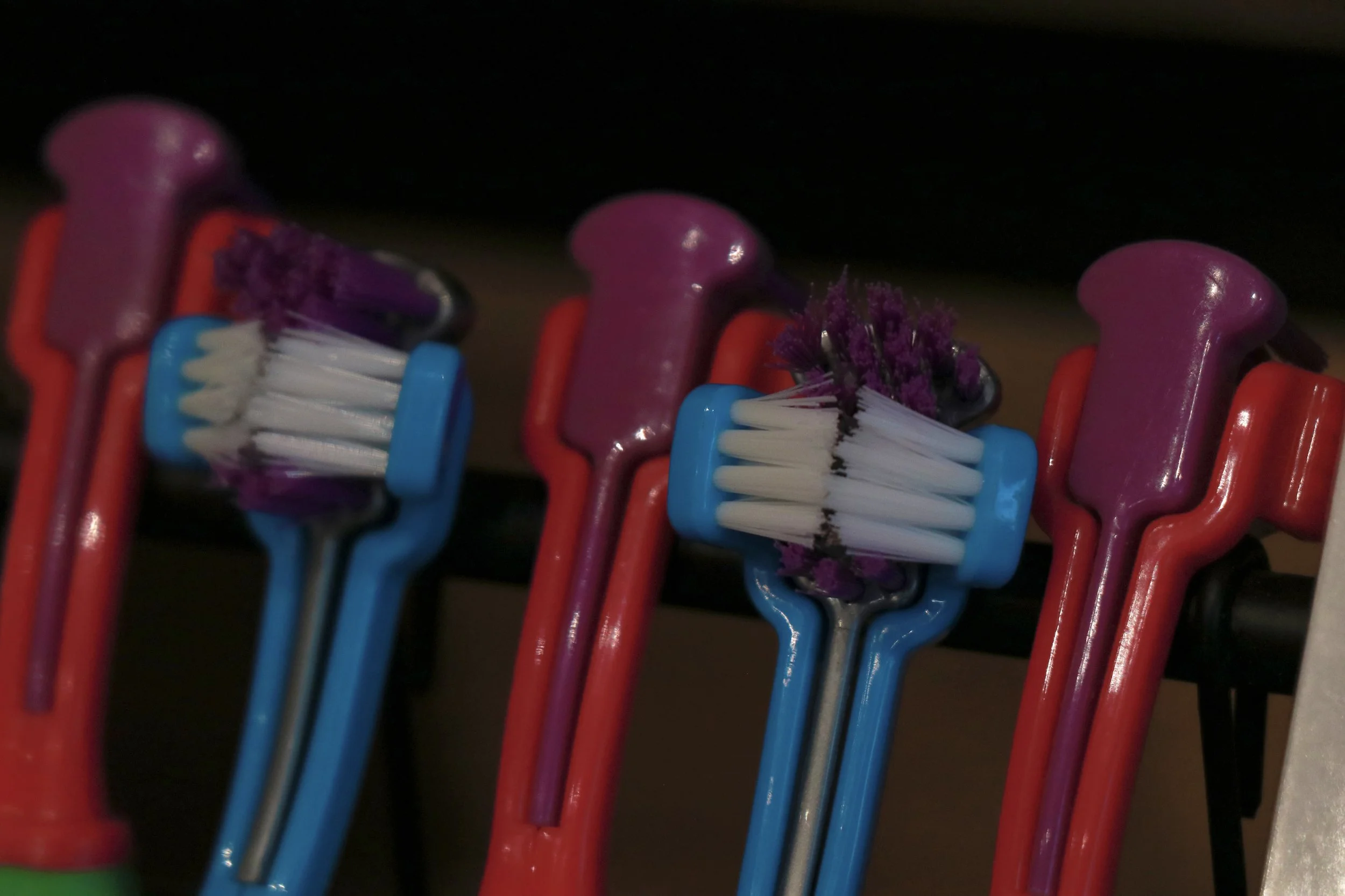 Colorful toothbrushes with purple, white, and black toothpaste on bristles, hanging on a black holder.