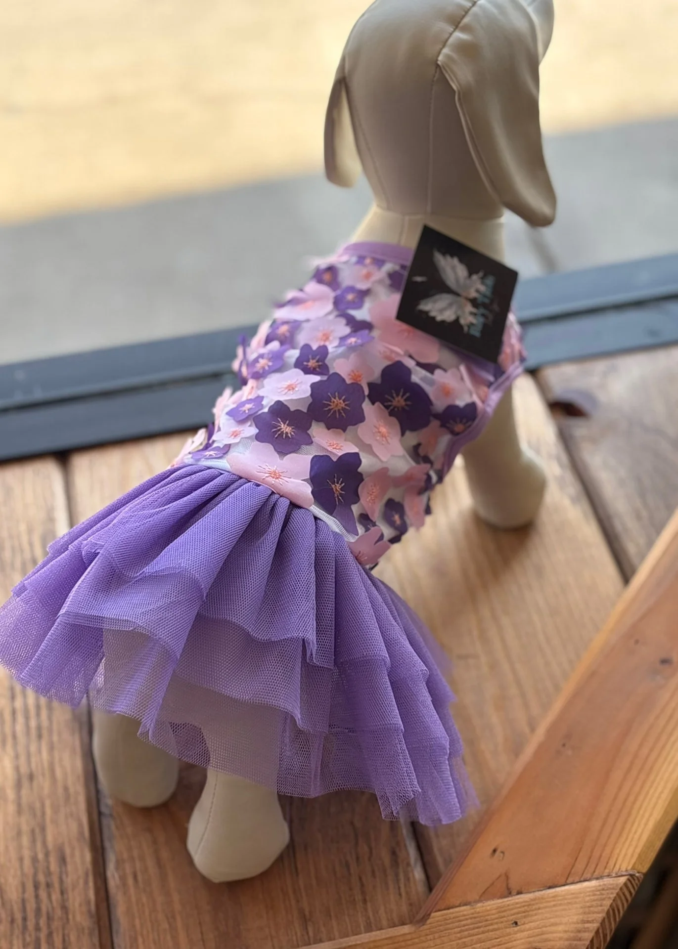 A mannequin dog dressed in a purple floral top with a purple tulle skirt, positioned on a wooden floor near a glass door.
