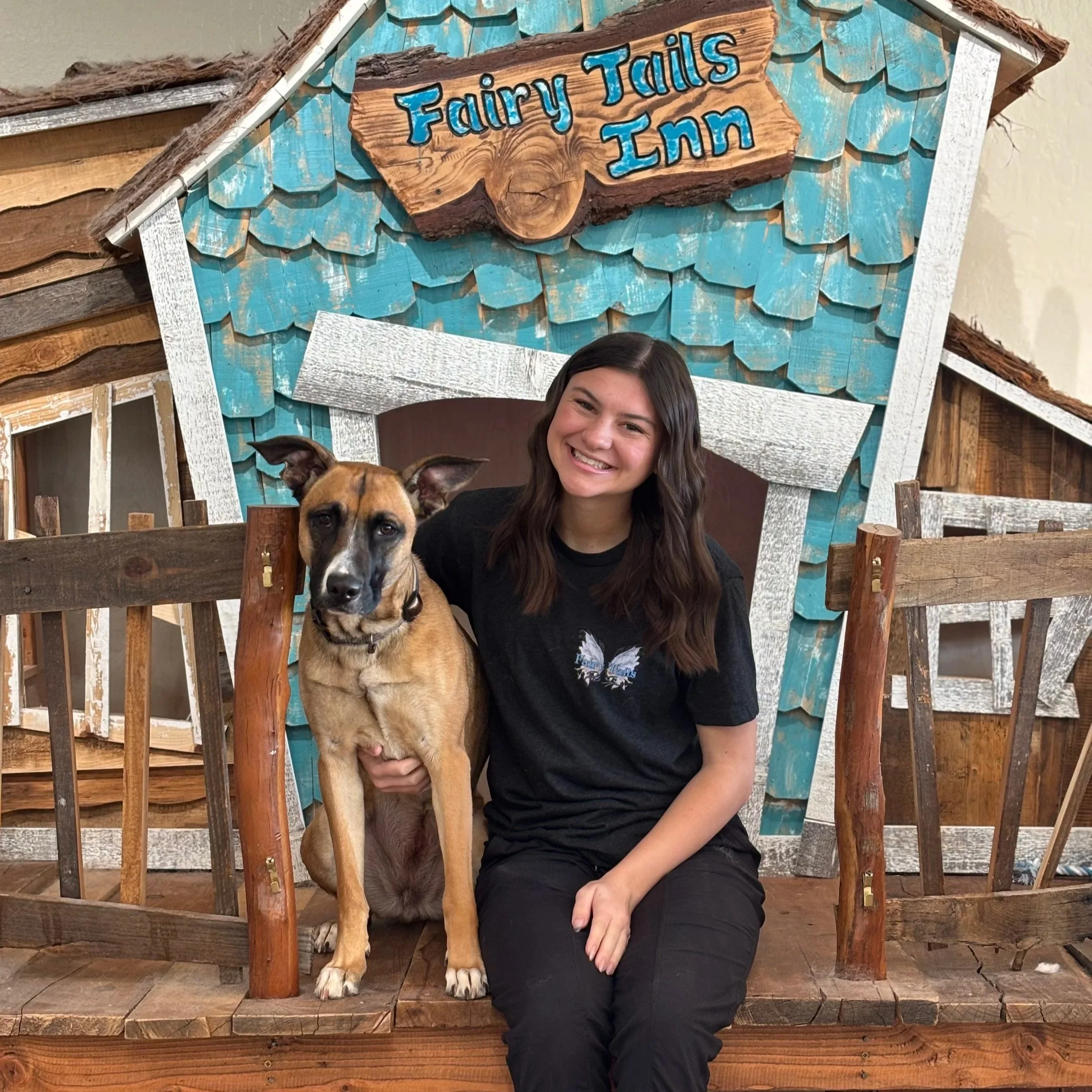 A young woman with long dark hair smiling while sitting next to a tan and white dog with a black mask and pointy ears, inside a wooden fairy tale themed structure with teal shingles and a sign that reads "Fairy Tales Inn".