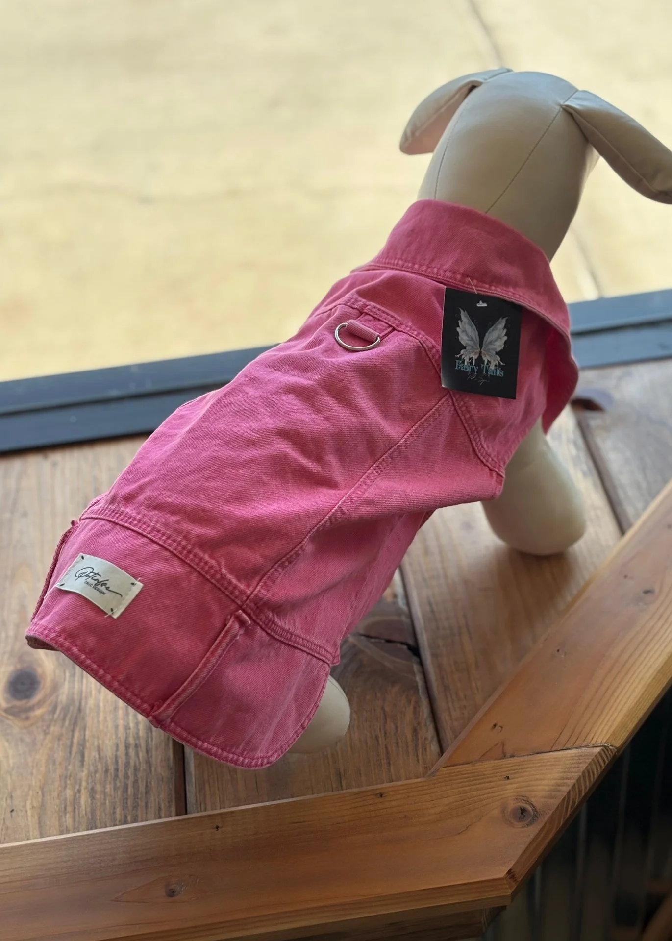 A mannequin dog wearing a pink denim jacket with a tag that has butterfly wings design, displayed on a wooden surface with a yellow background.