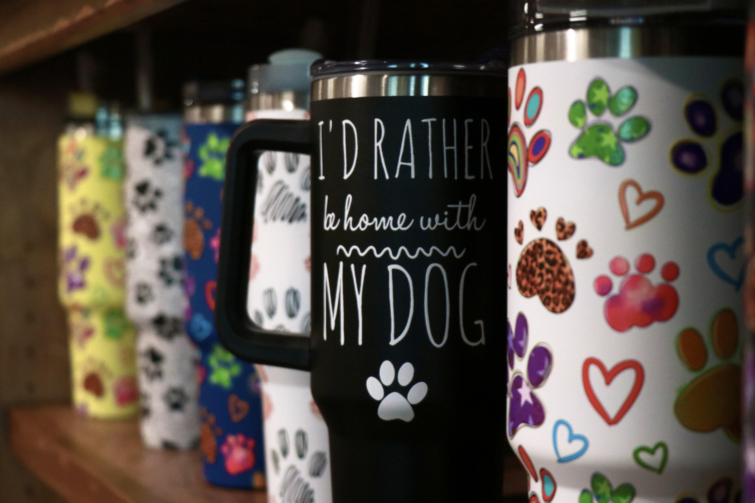 Colorful collection of pet-themed mugs with paw prints, hearts, and playful designs on a wooden shelf.
