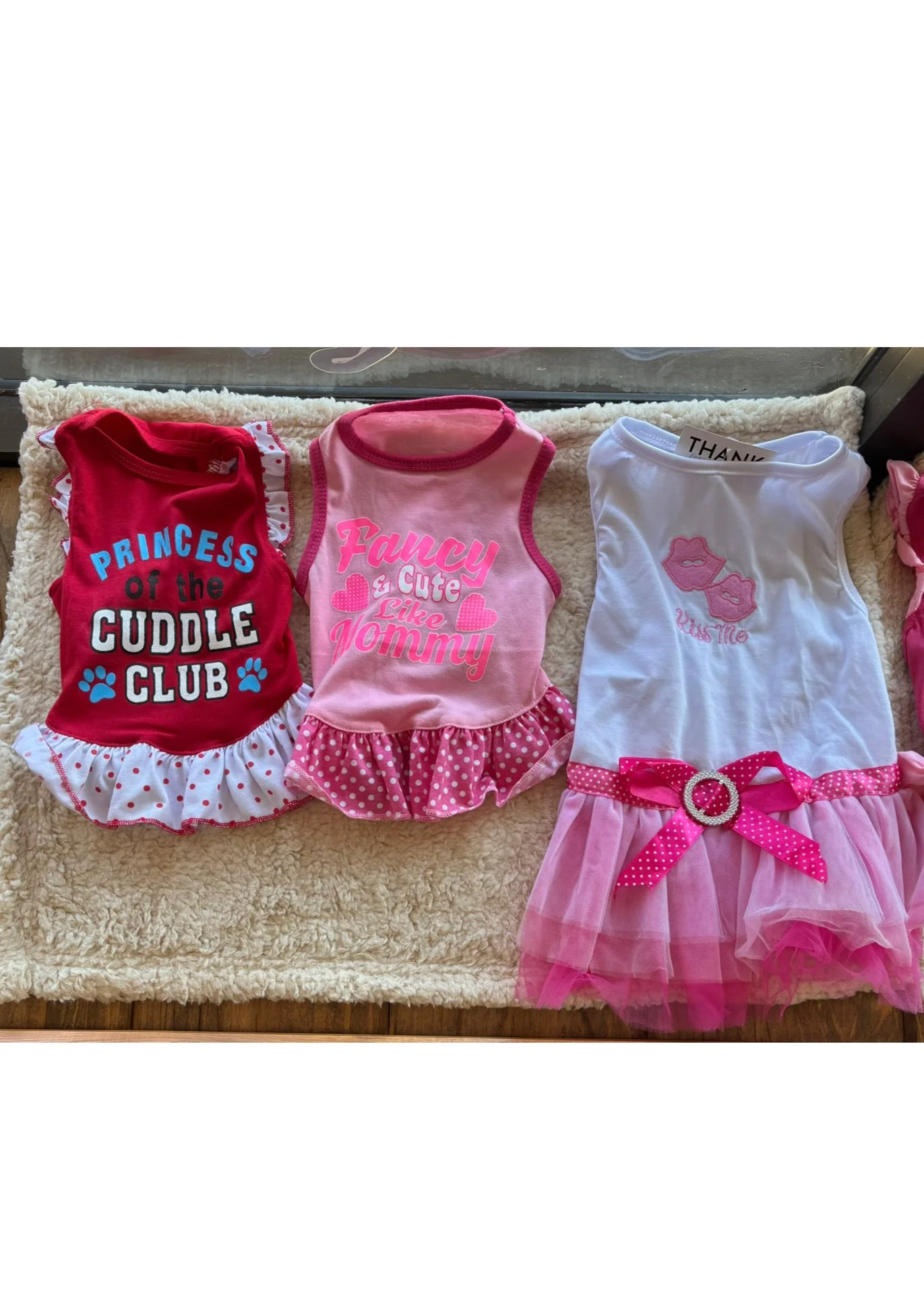 Three children's sleeveless dresses with pink, red, and white themes and playful text and designs on a beige fuzzy surface.