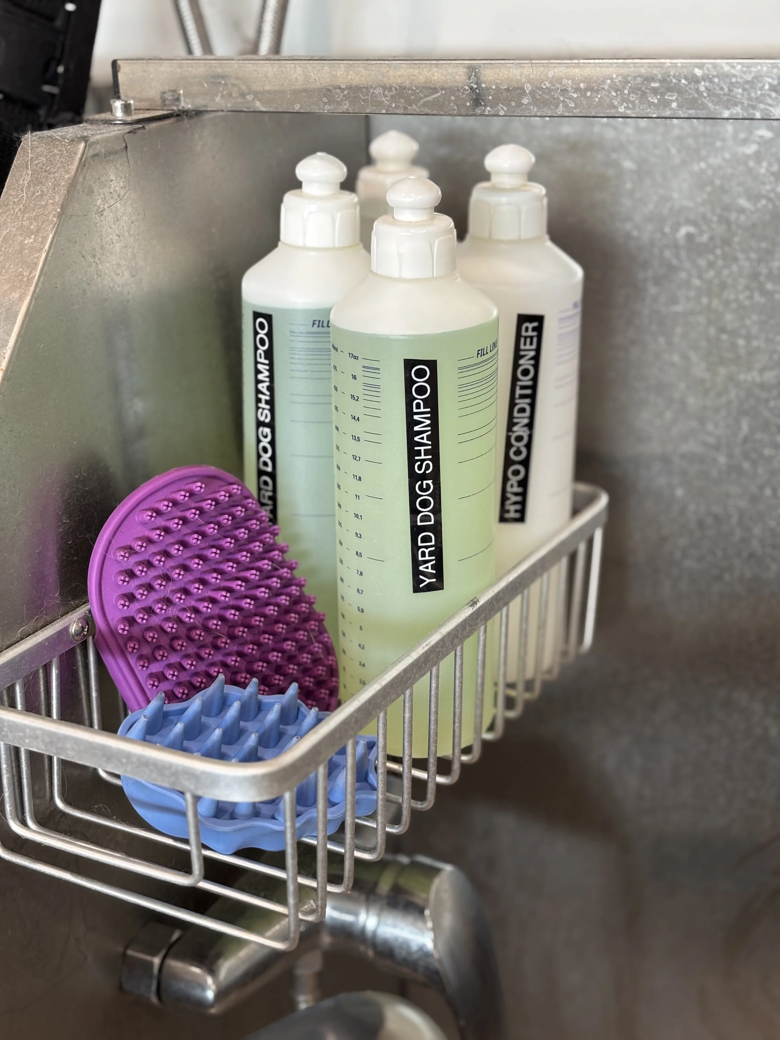 A metal shower caddy containing bottles of yard dog shampoo and conditioner, along with purple and blue silicone grooming brushes.