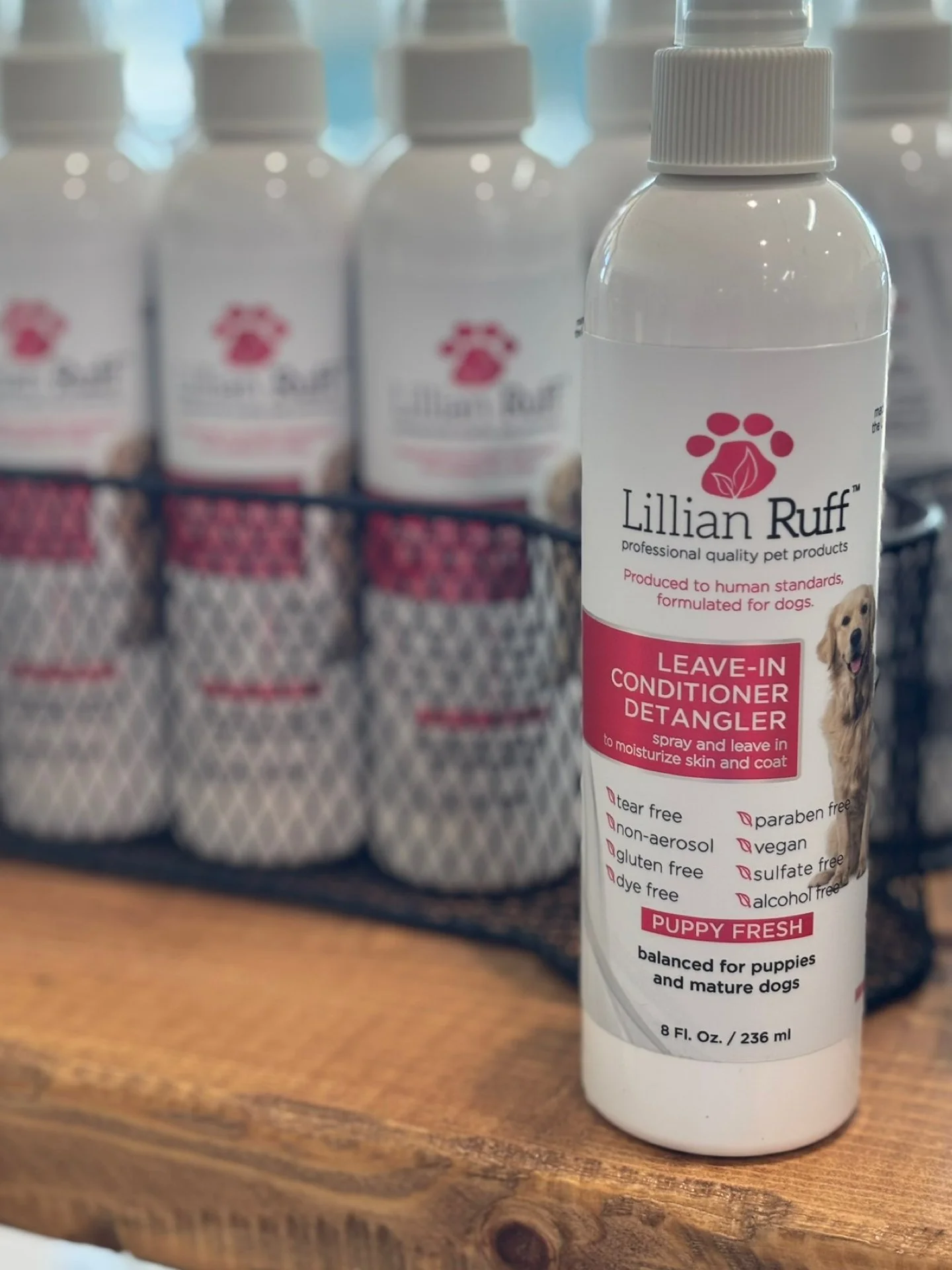 A spray bottle of Lillian Ruff leave-in conditioner detangler for dogs, positioned on a wooden surface with other identical bottles in the background. The label indicates it is for puppies and mature dogs, and features a picture of a dog.