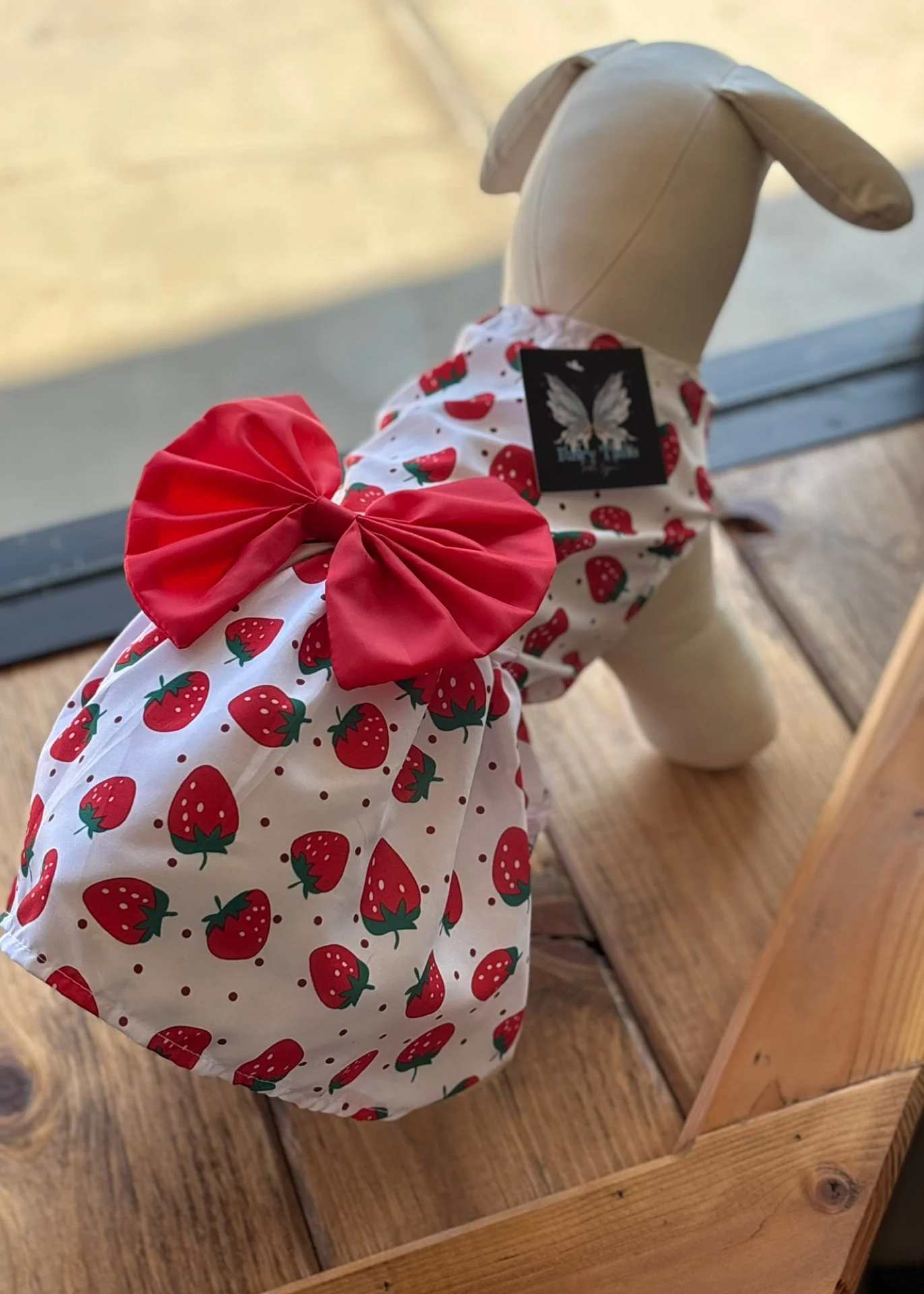 A stuffed dog toy dressed in a strawberry-patterned dress with a large red bow on the back, sitting on a wooden surface near a window.
