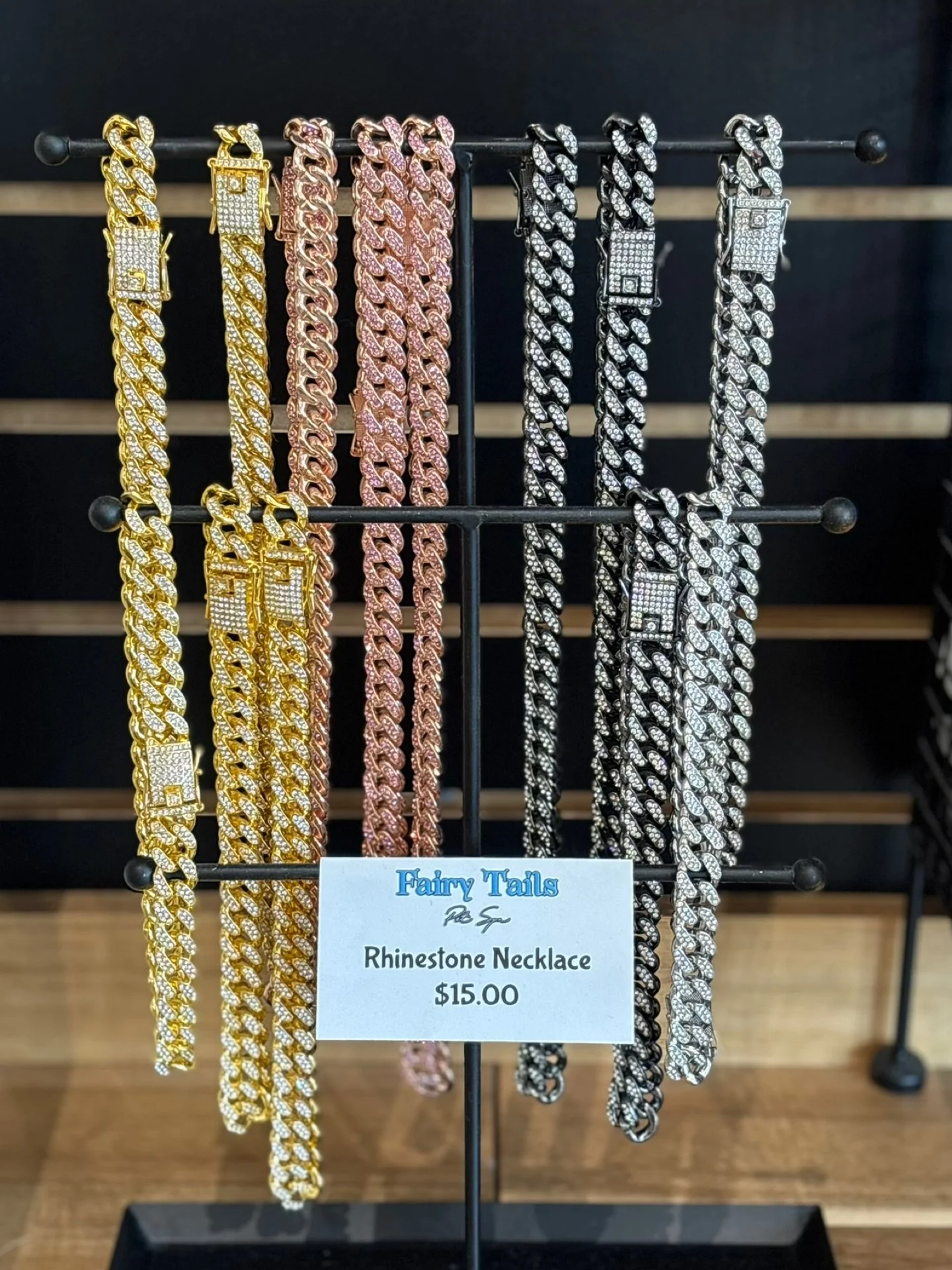 Collection of gold, rose gold, silver, and black rhinestone jewelry necklaces displayed on a black stand with a sign reading 'Fairy Tails Rhinestone Necklace $15.00'.