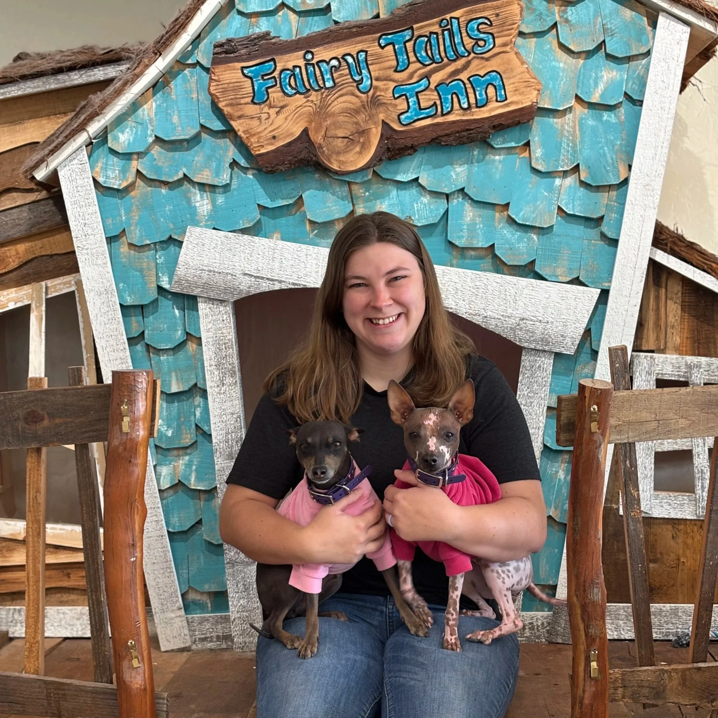 A woman sitting on a wooden chair holding two dogs, with a mural behind her that reads 'Fairy Tails Inn'.