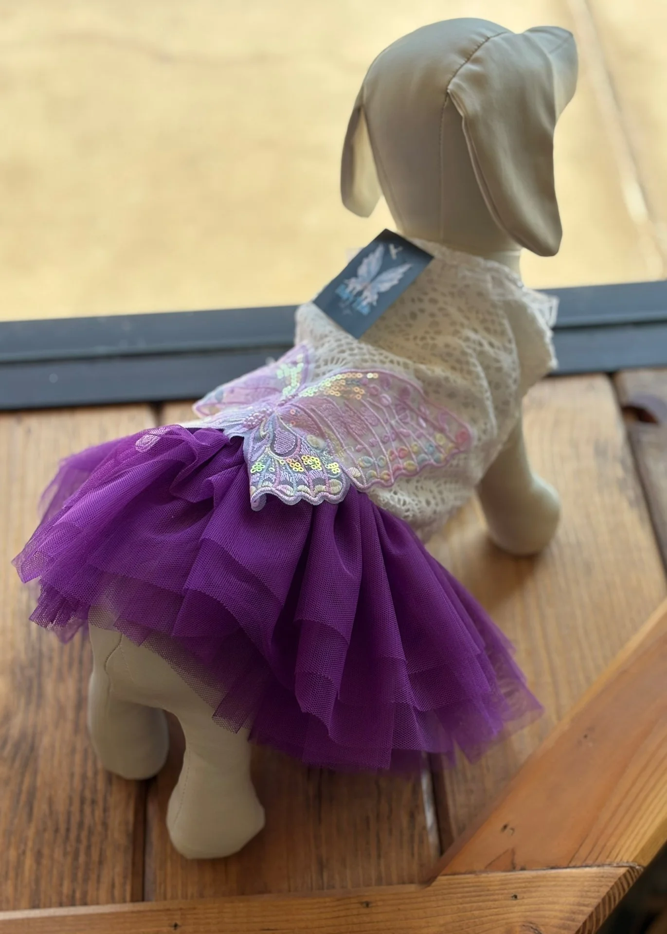 A plush dog wearing a purple tutu and a lace top, displayed on a wooden surface near a window.