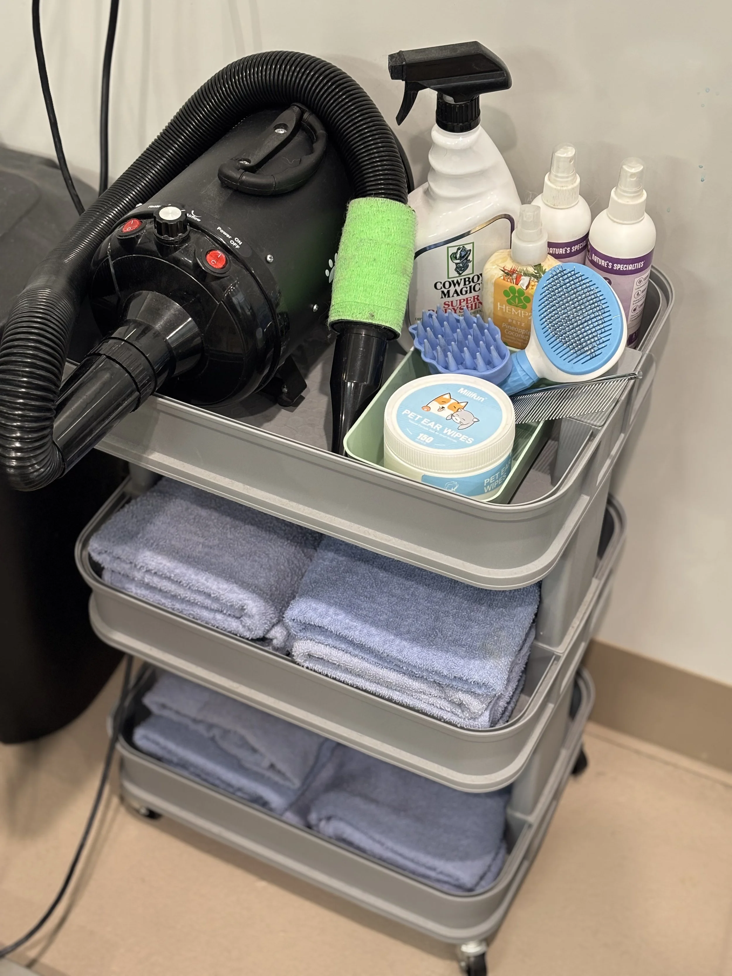 A rolling cart with cleaning supplies, including towels, pet wipes, spray bottles, a vacuum cleaner, and grooming brushes.
