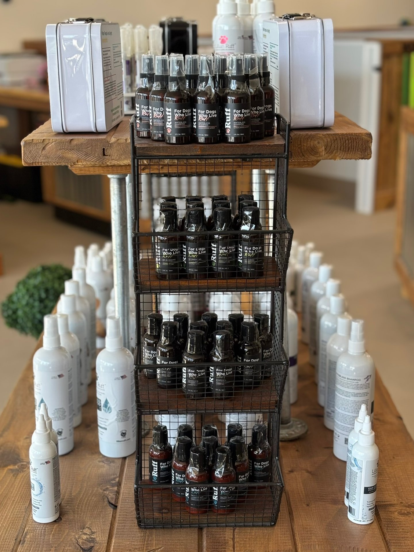Display of pet care products on a wooden table, including white spray bottles and small dark spray bottles labeled for dogs, organized in a black wire rack with multiple shelves.
