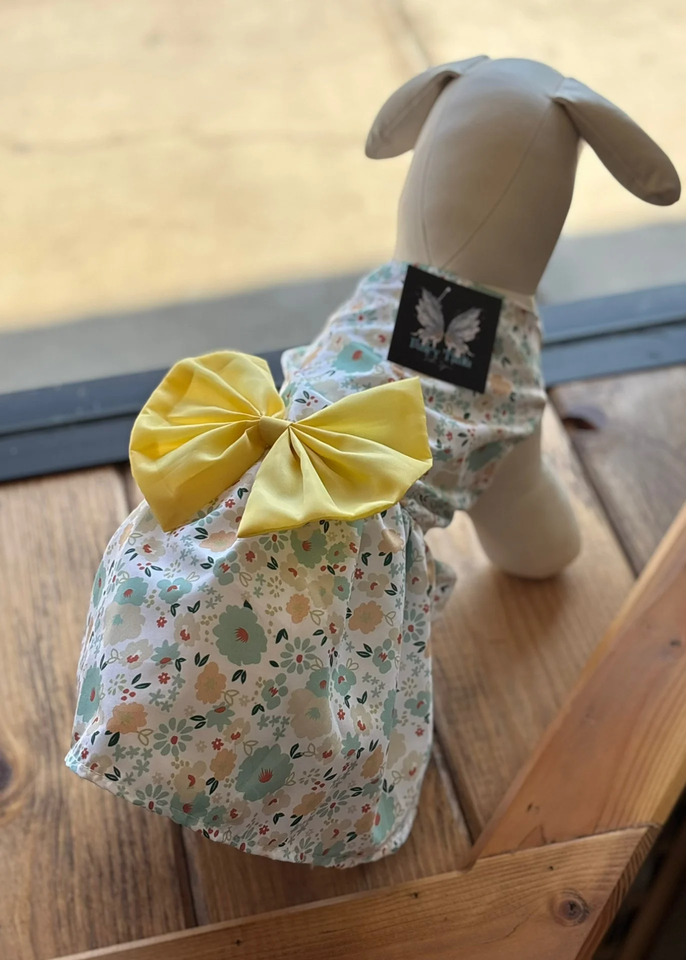 A stuffed dog toy dressed in a floral dress with a yellow bow, sitting on a wooden surface near a window.