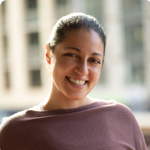 A woman smiling with sunlight on her face, out of focus city windows in the background.