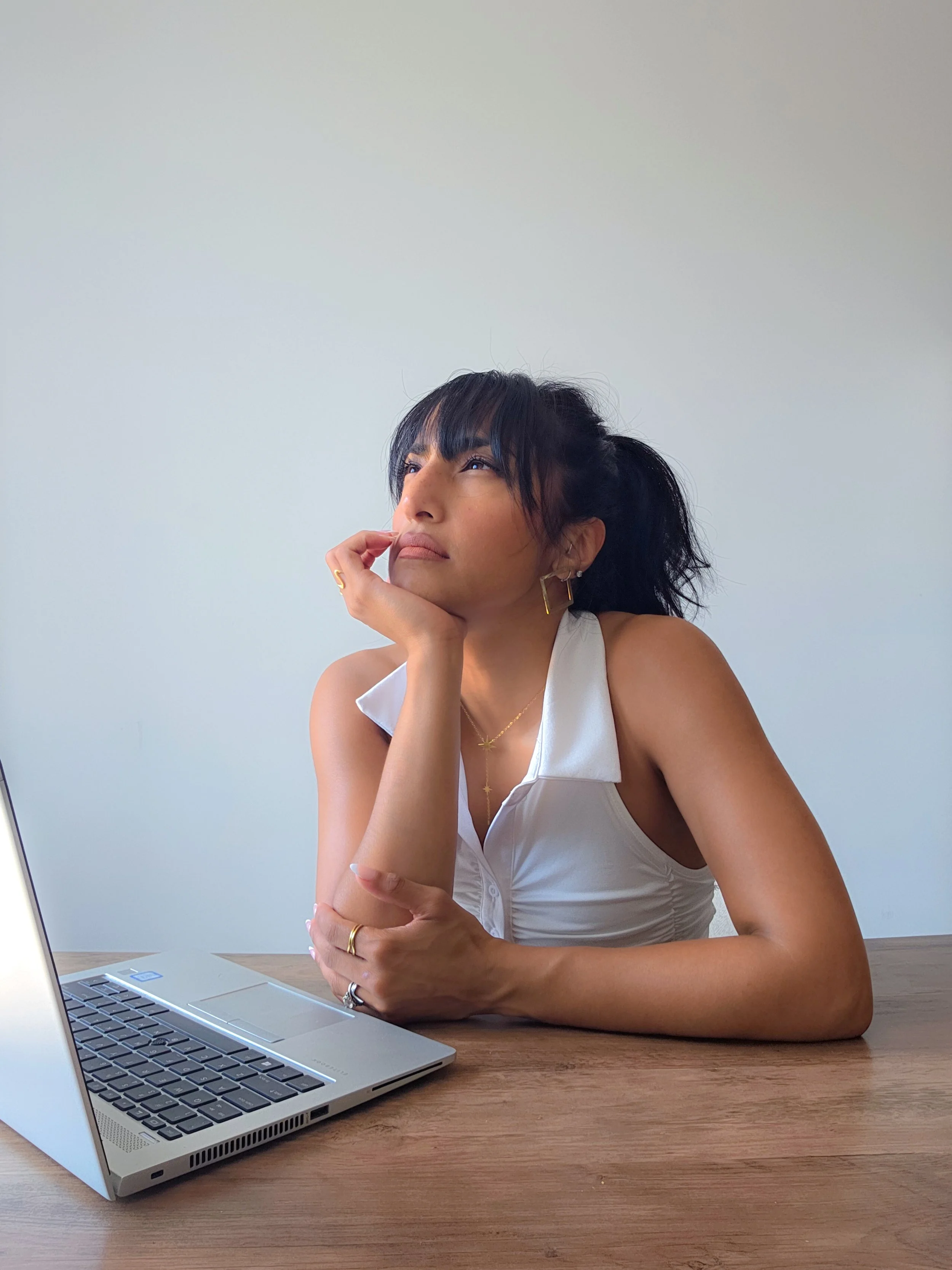 A woman with dark hair and gold jewelry sitting at a wooden table with her chin resting on her hand and a laptop in front of her, looking thoughtfully to the side against a plain white wall.