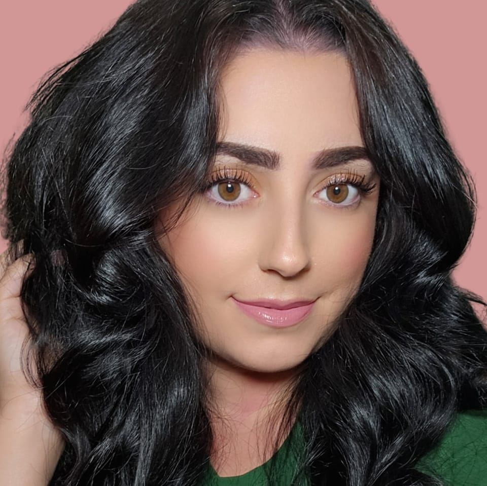 A woman with long, black, wavy hair, brown eyes, and fair skin, smiling softly against a pink background.