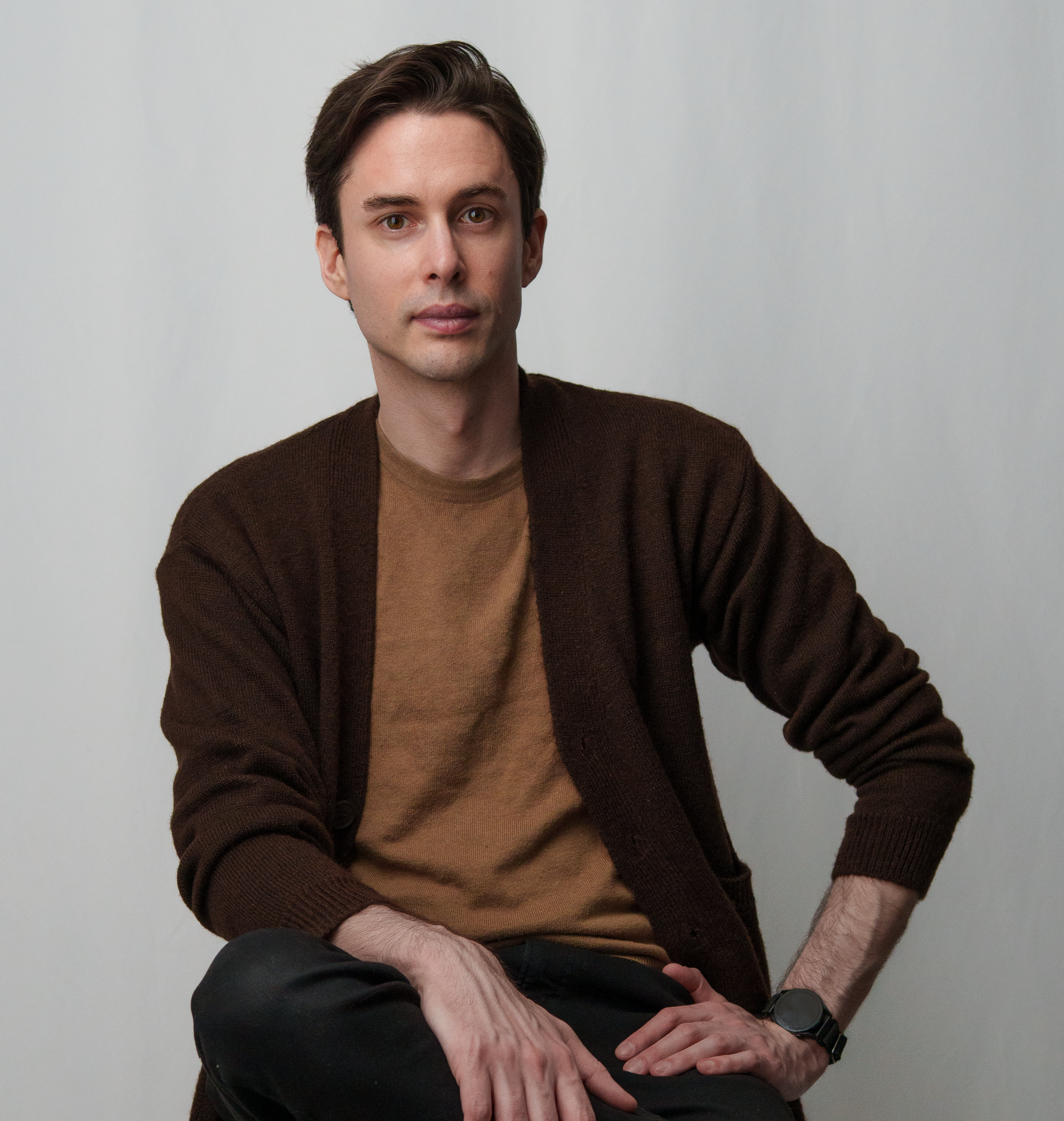 A young man with brown hair, wearing a brown cardigan over a tan shirt, sitting against a plain light gray background, looking directly at the camera.
