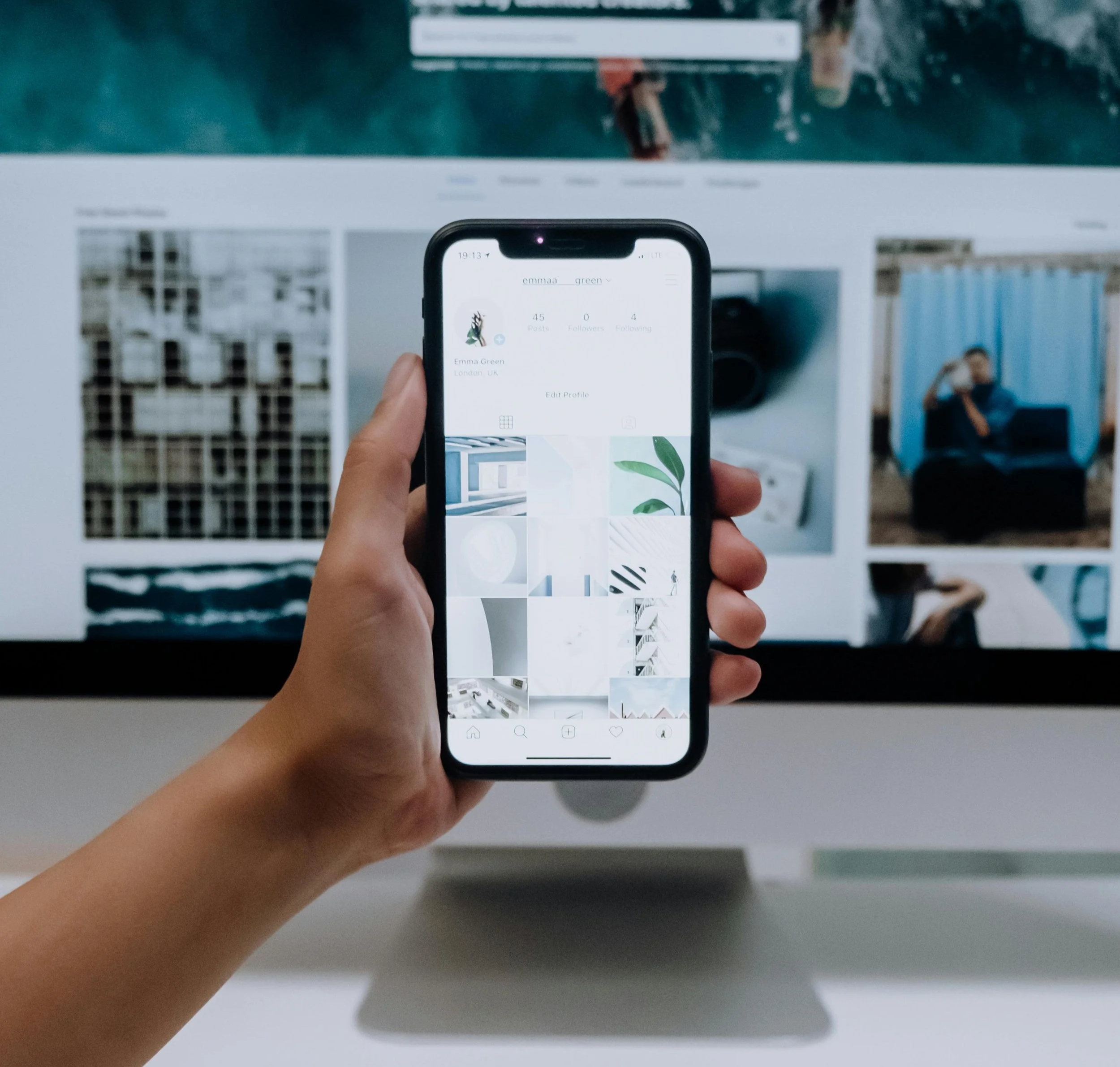 Person holding a smartphone in front of a computer screen displaying a photo gallery or social media profile with various pictures.