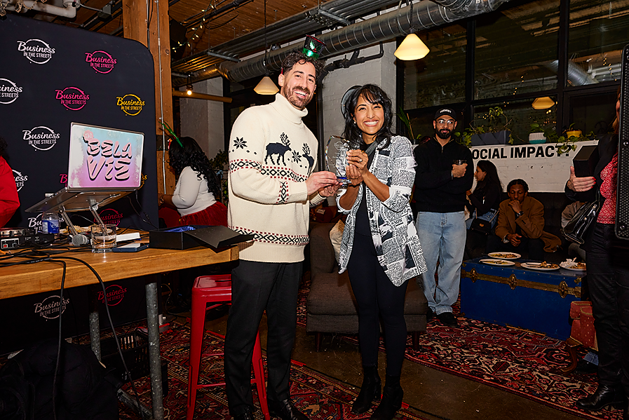 Two people smiling and holding an award at a social event in a decorated indoor venue with a sign that says 'Social Impact' and a DJ setup.