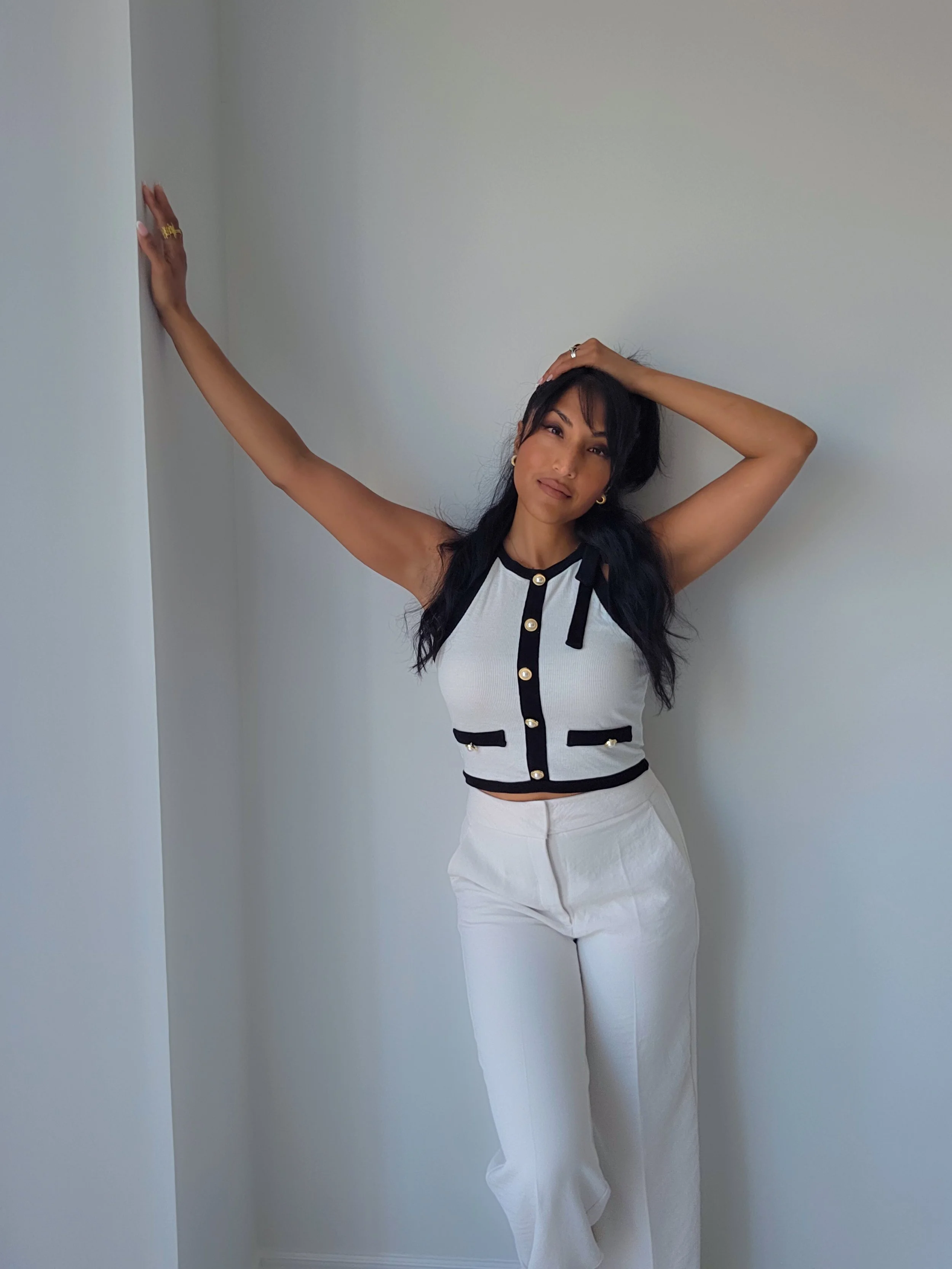 A woman with long dark hair wearing a sleeveless white top with black trim and gold buttons, and white pants, standing against a plain white wall with one arm raised and resting against the wall, and the other hand on her head, looking directly at the camera.