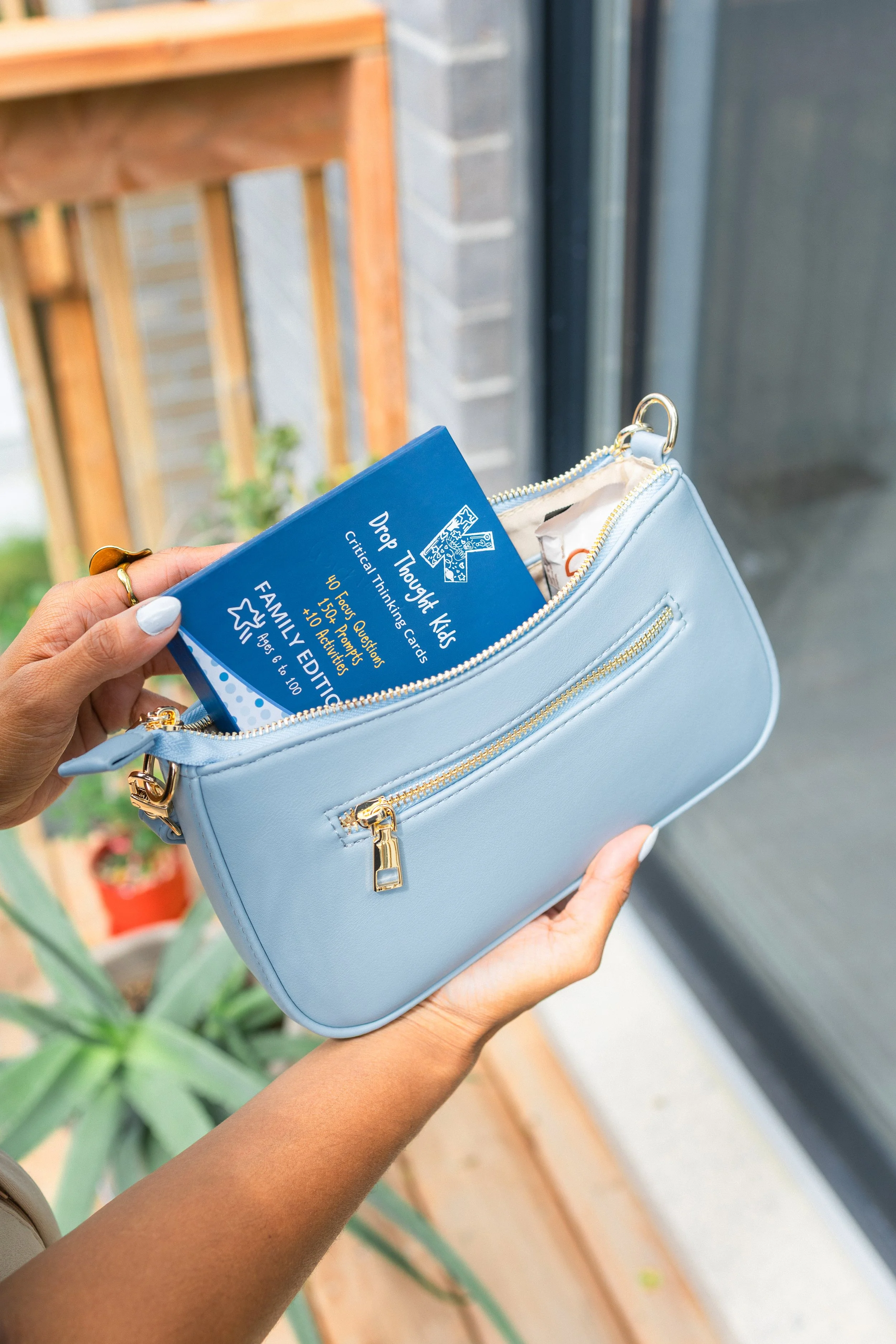 Person holding a light blue purse with gold zipper, placing a blue card inside, near a window with plants and brick wall visible outside.