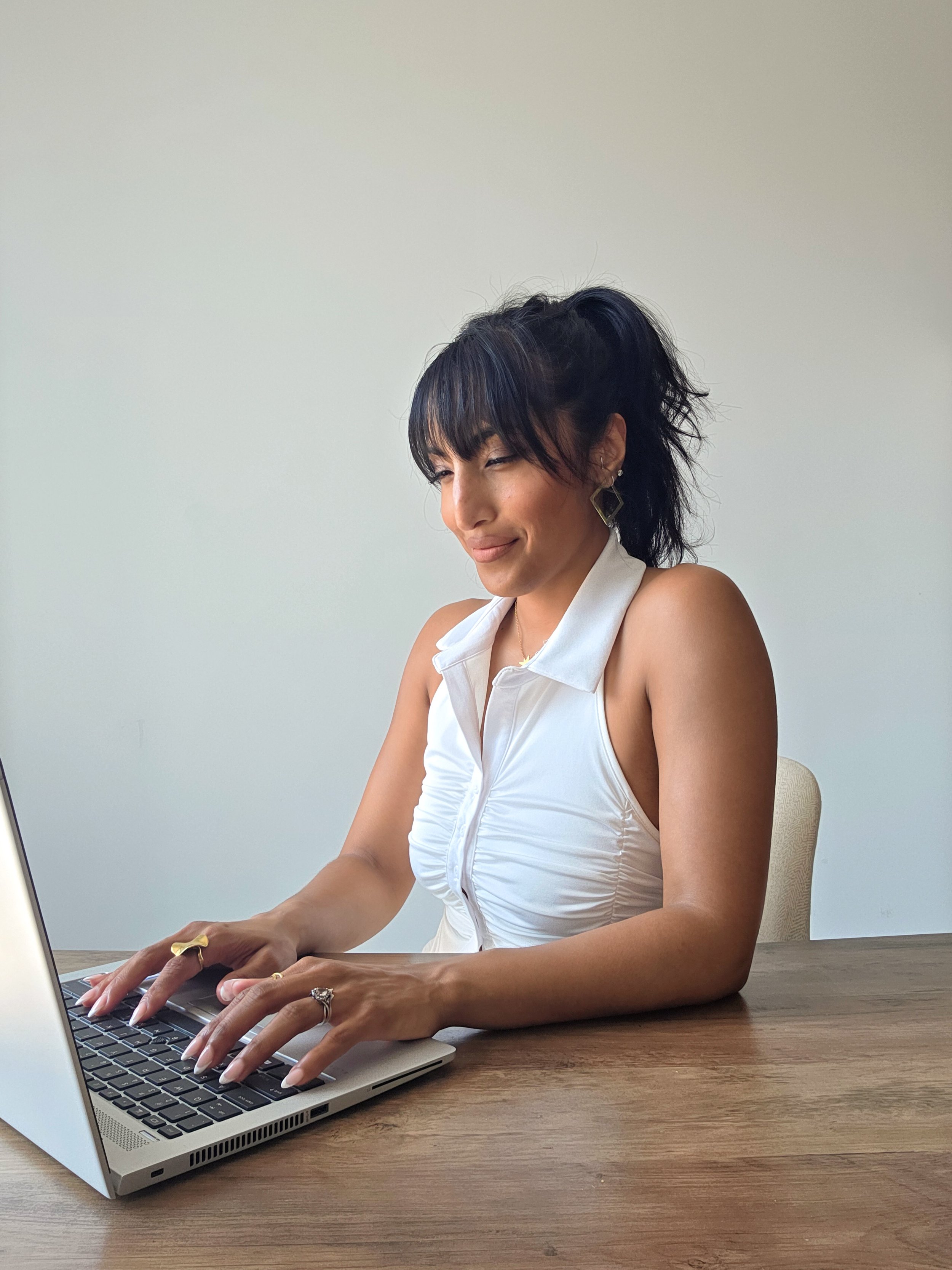 Woman with dark hair styled in a high ponytail, wearing a sleeveless white top, working on a laptop at a wooden table.