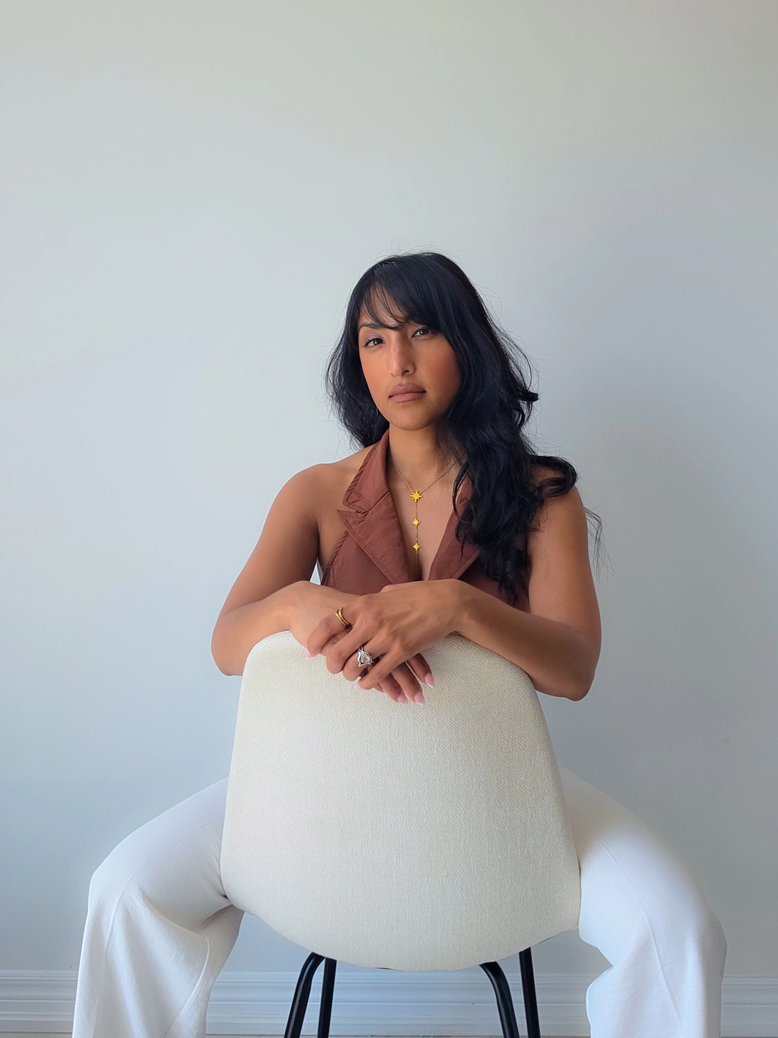 A woman with long dark hair and tan skin is sitting in a white chair, wearing a brown sleeveless top, white pants, and gold jewelry, with a plain light-colored wall in the background.
