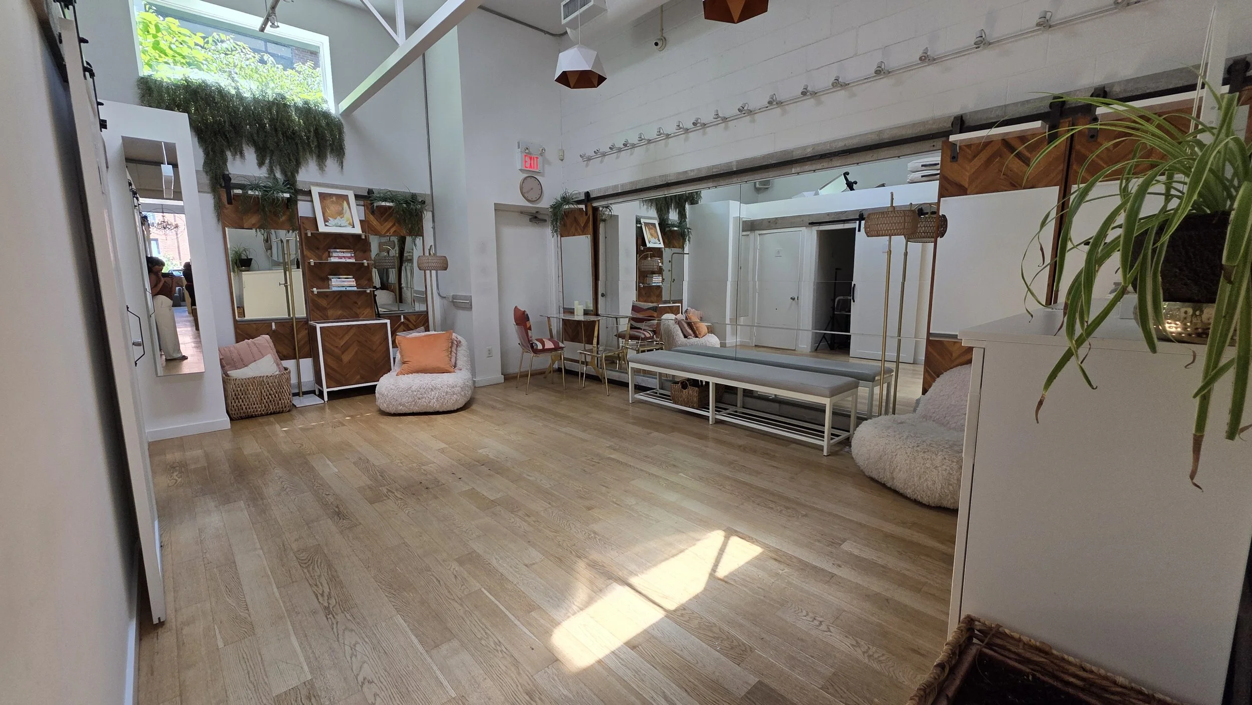 Interior of a modern, well-lit room with wooden flooring, featuring a seating area with plush chairs and cushions, bookshelves with plants, and large windows letting in natural light.